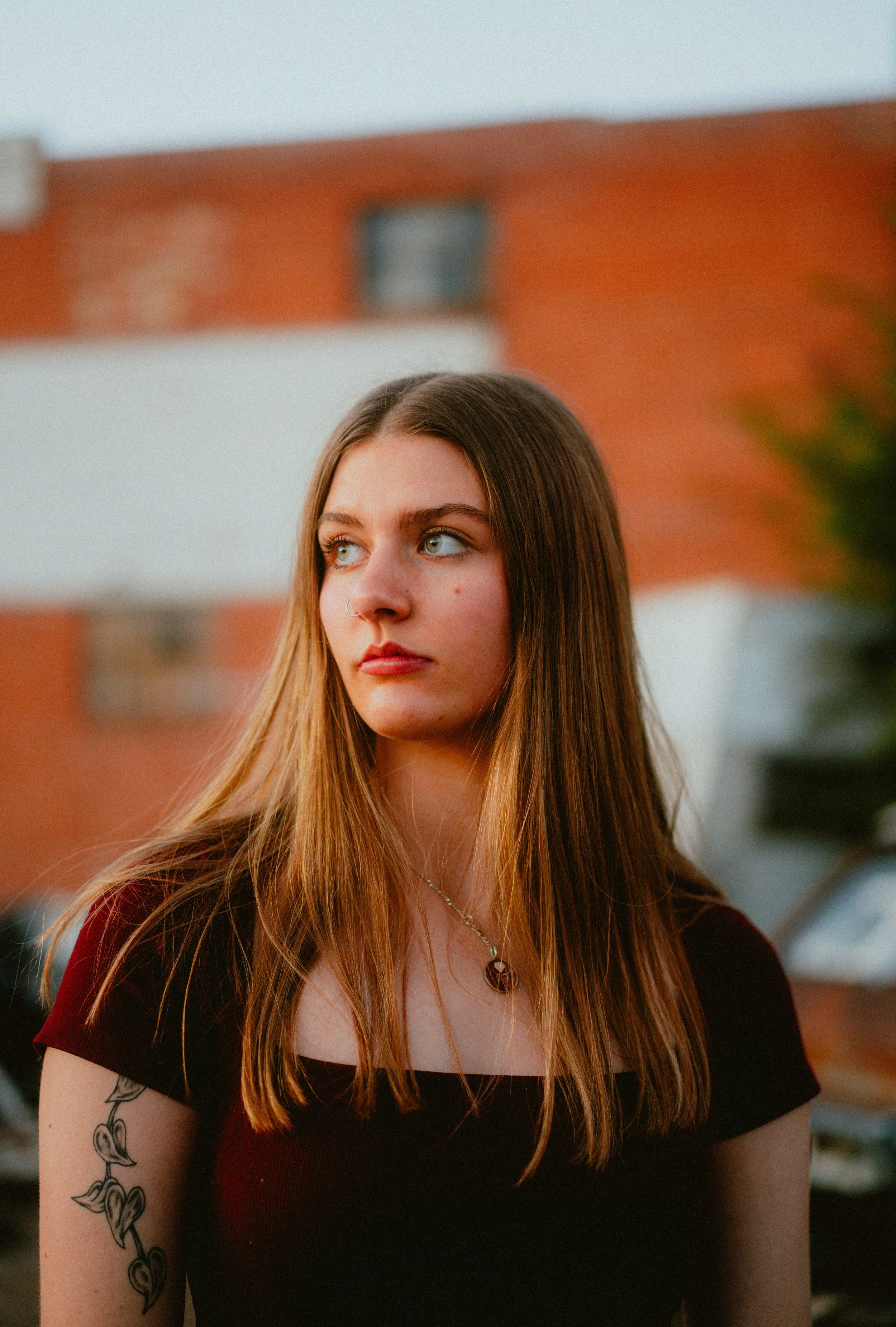 Young woman with long brown hair, blue eyes, and a nose piercing, wearing a dark red and black top, gold necklace, and a tattoo on her upper arm, standing outdoors with a blurred background of a brick building and sky.