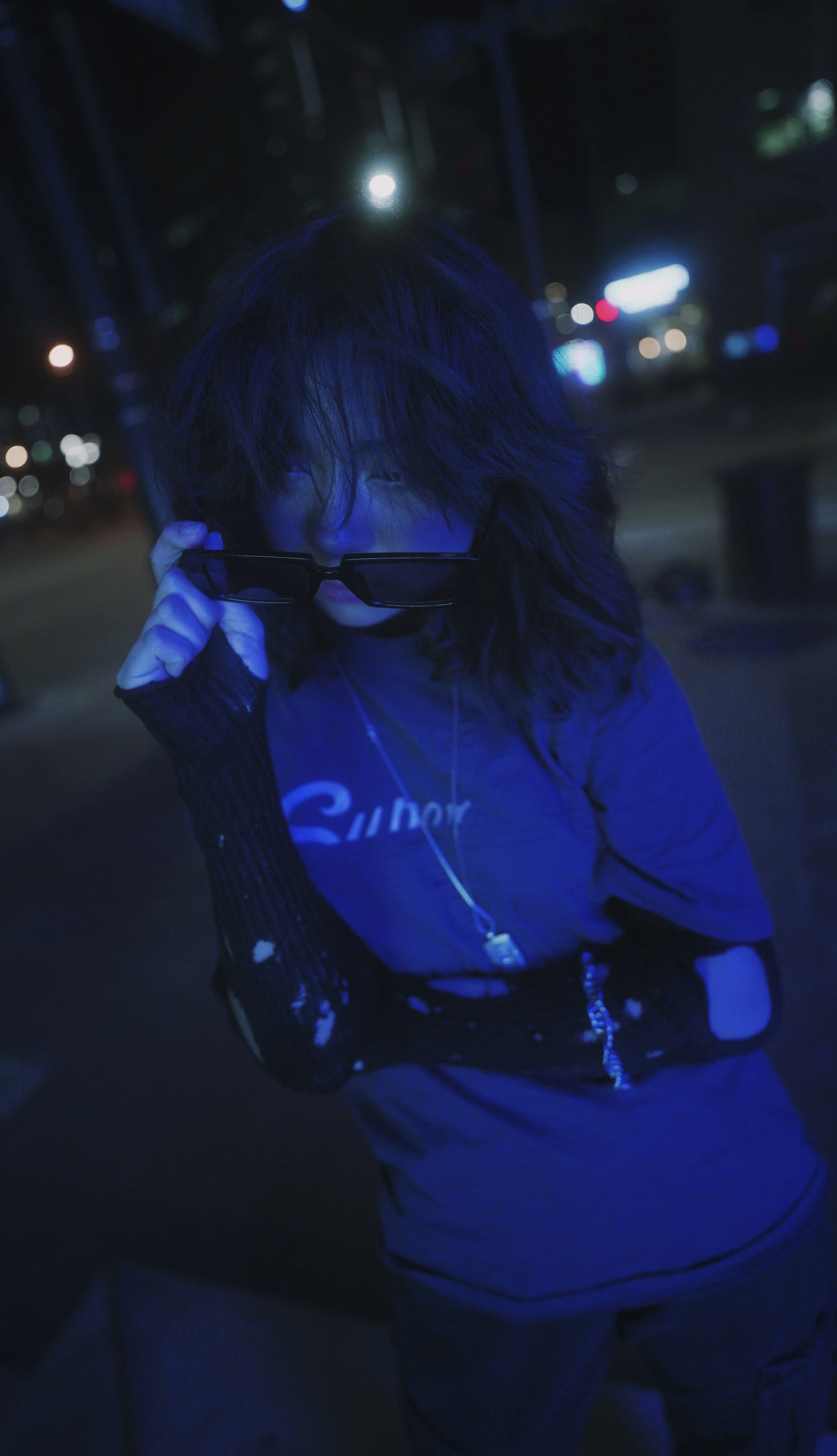 A person with dark hair wearing glasses and a blue shirt, adjusting their glasses while standing outdoors at night with city lights in the background.