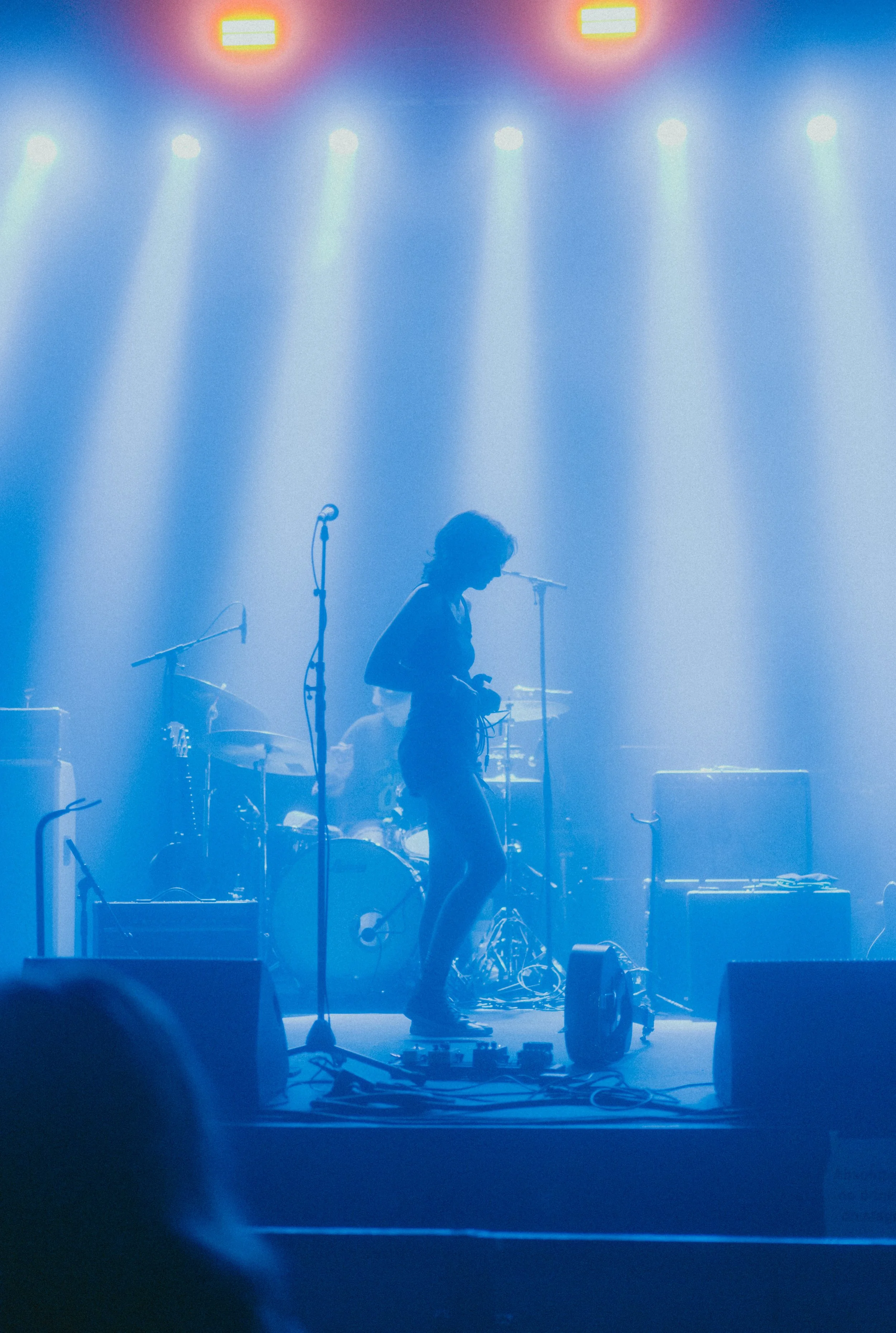 Concert stage with blue lighting, a person adjusting equipment, microphones, drums, and musical instruments, illuminated by stage lights.