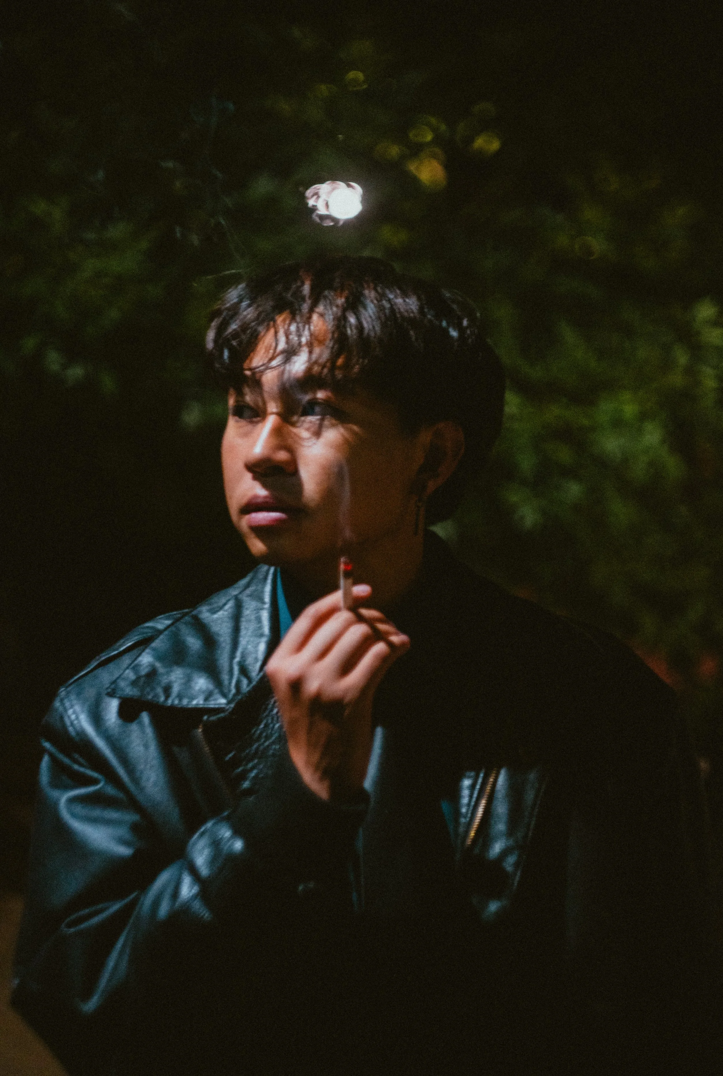 A young man with dark, short hair and a piercing in his ear is holding a lit cigarette in his right hand, wearing a shiny black leather jacket, standing outdoors at night with a dark, leafy background.
