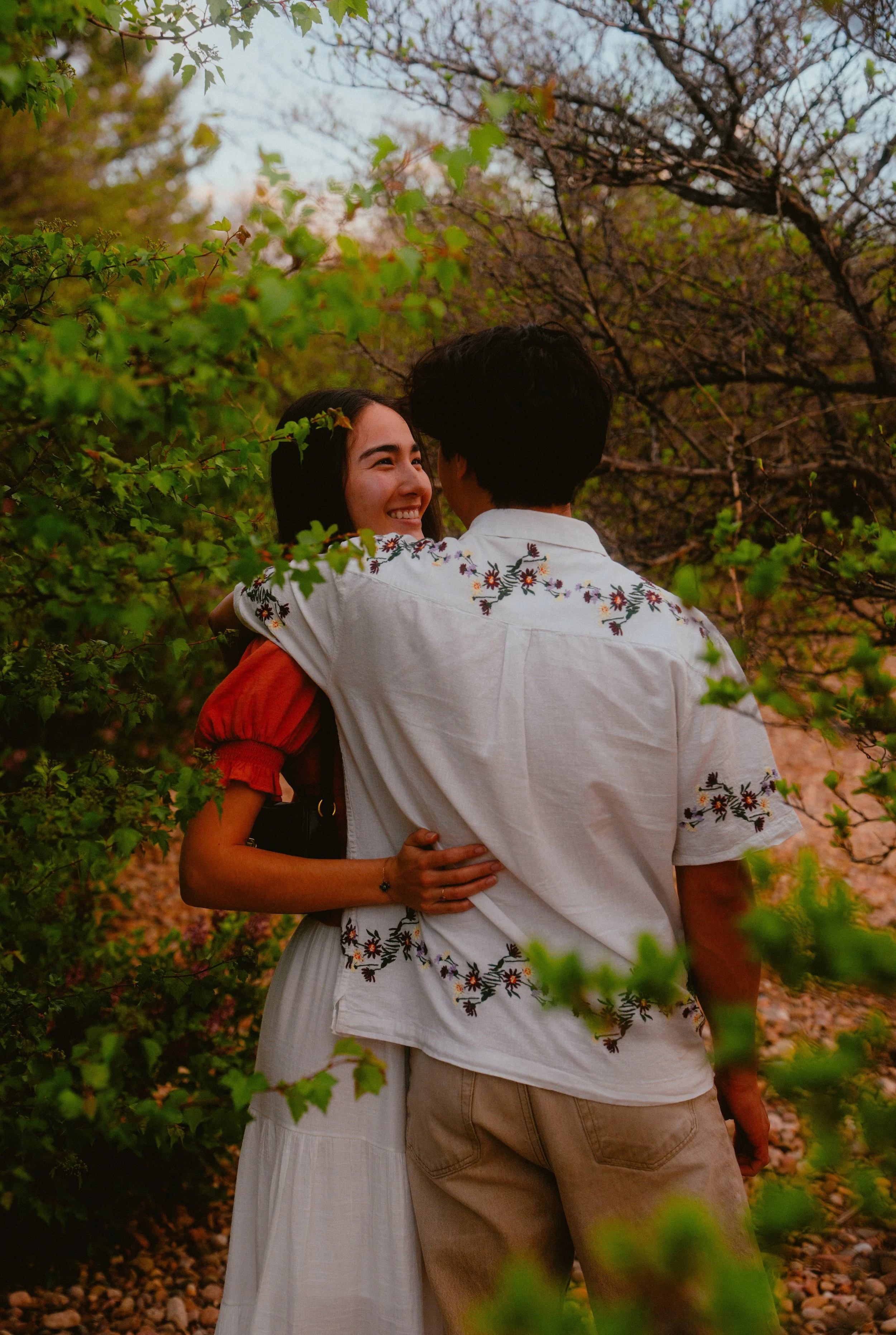 A couple embraces outdoors among trees, with the woman smiling at the man. The woman wears a white dress with floral embroidery, and the man wears a white shirt with floral embroidery on the back and beige pants.