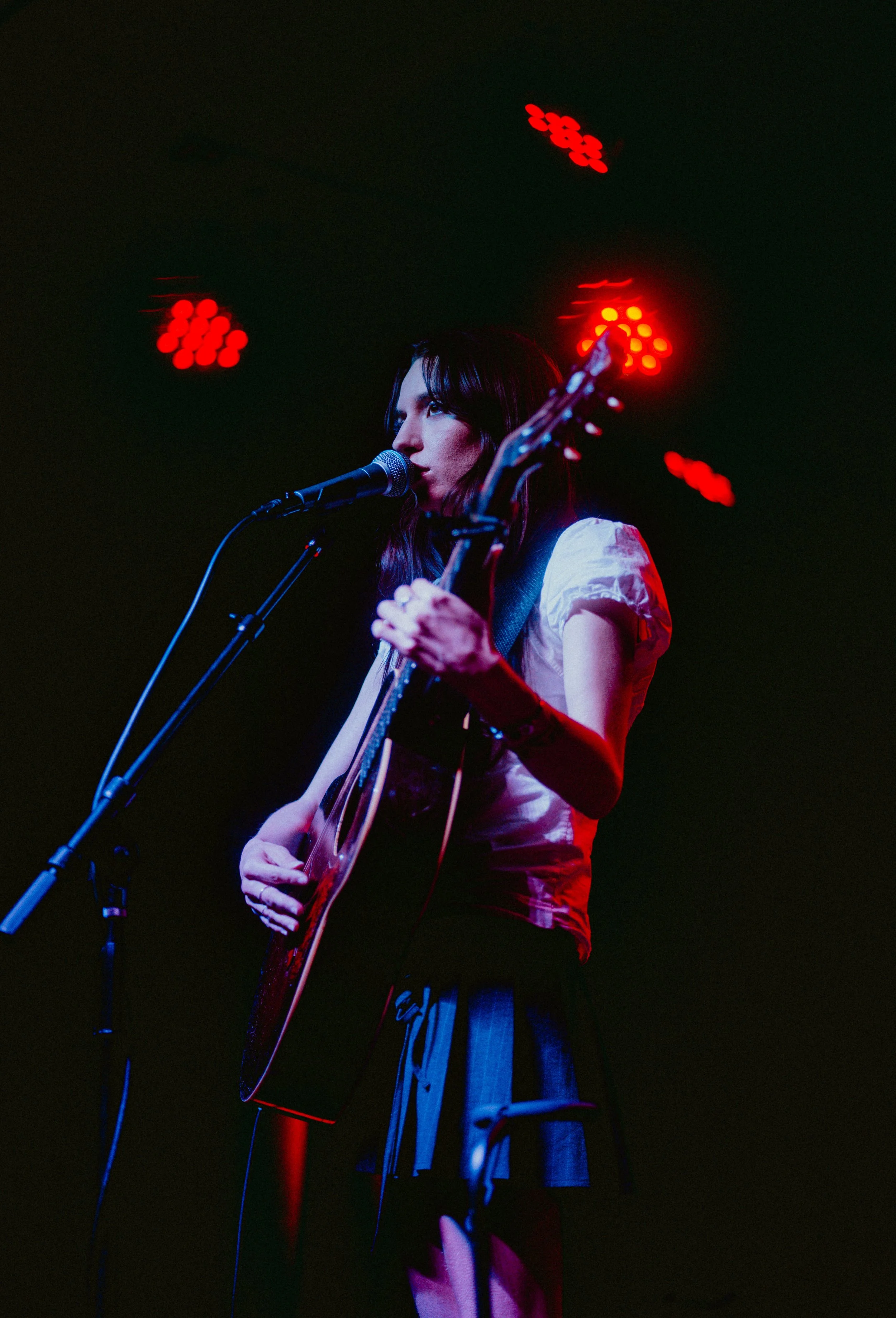 A woman with dark hair is singing into a microphone while playing an acoustic guitar on stage. Red stage lights are illuminating the dark background.