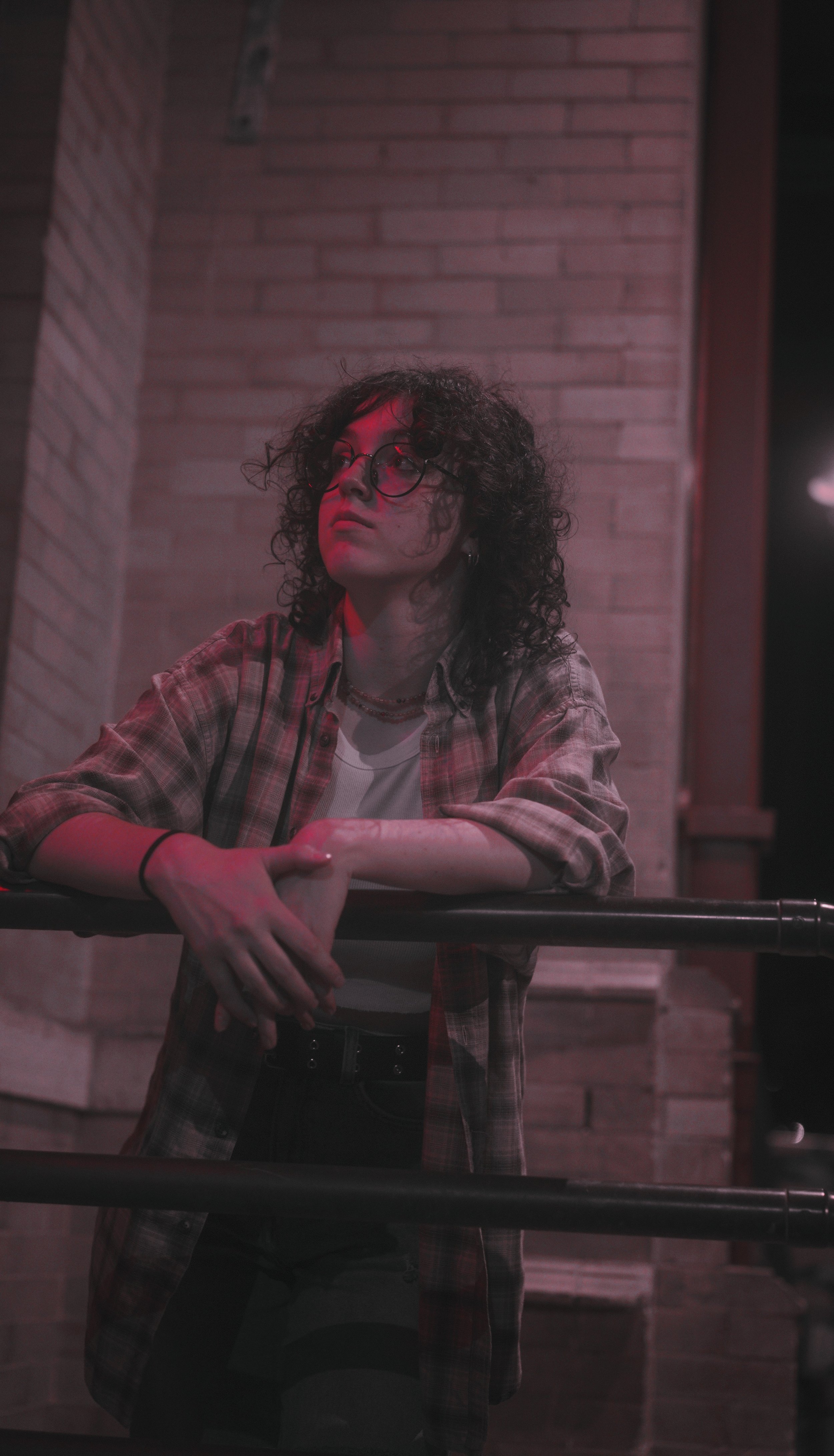 A young woman with curly hair, wearing glasses, a plaid shirt, and multiple necklaces, leaning on a railing against a brick wall background in a dimly lit setting.