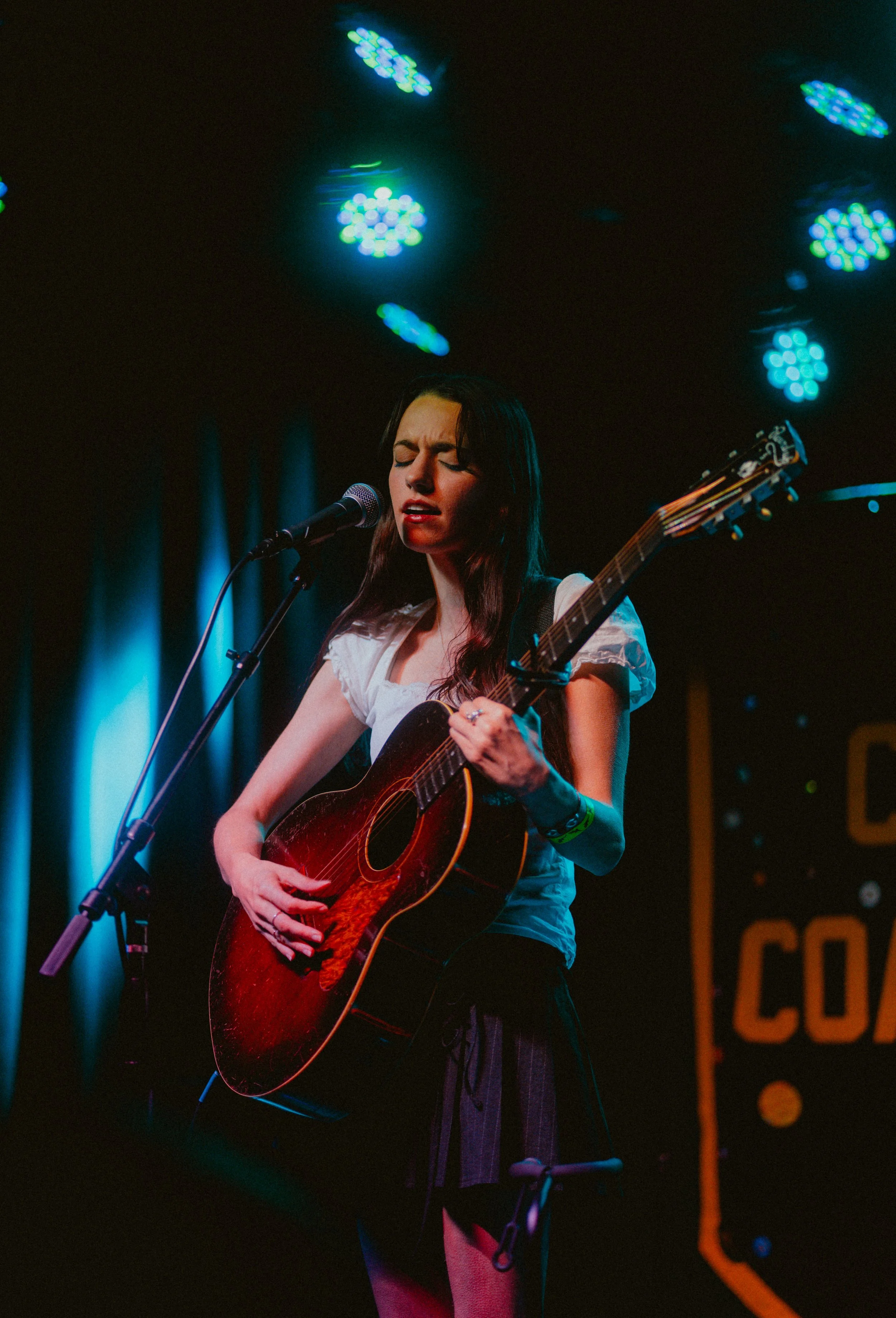 A young woman with long dark hair singing into a microphone while playing an acoustic guitar on stage, with colorful stage lights overhead.