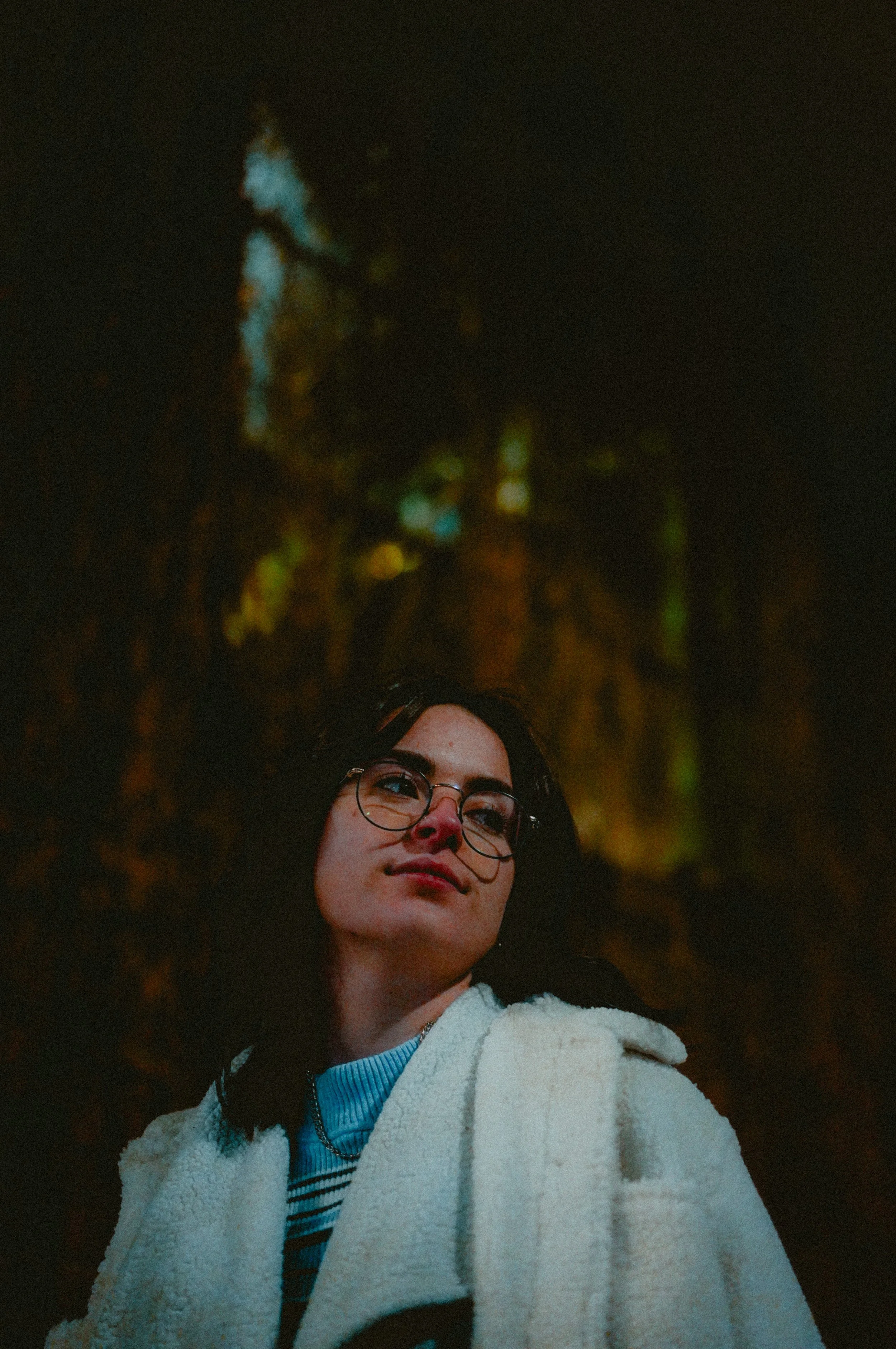 A woman with glasses and dark hair looking off to the side, wearing a cream-colored fluffy coat and a blue striped sweater underneath, with a blurred background of colorful lights.