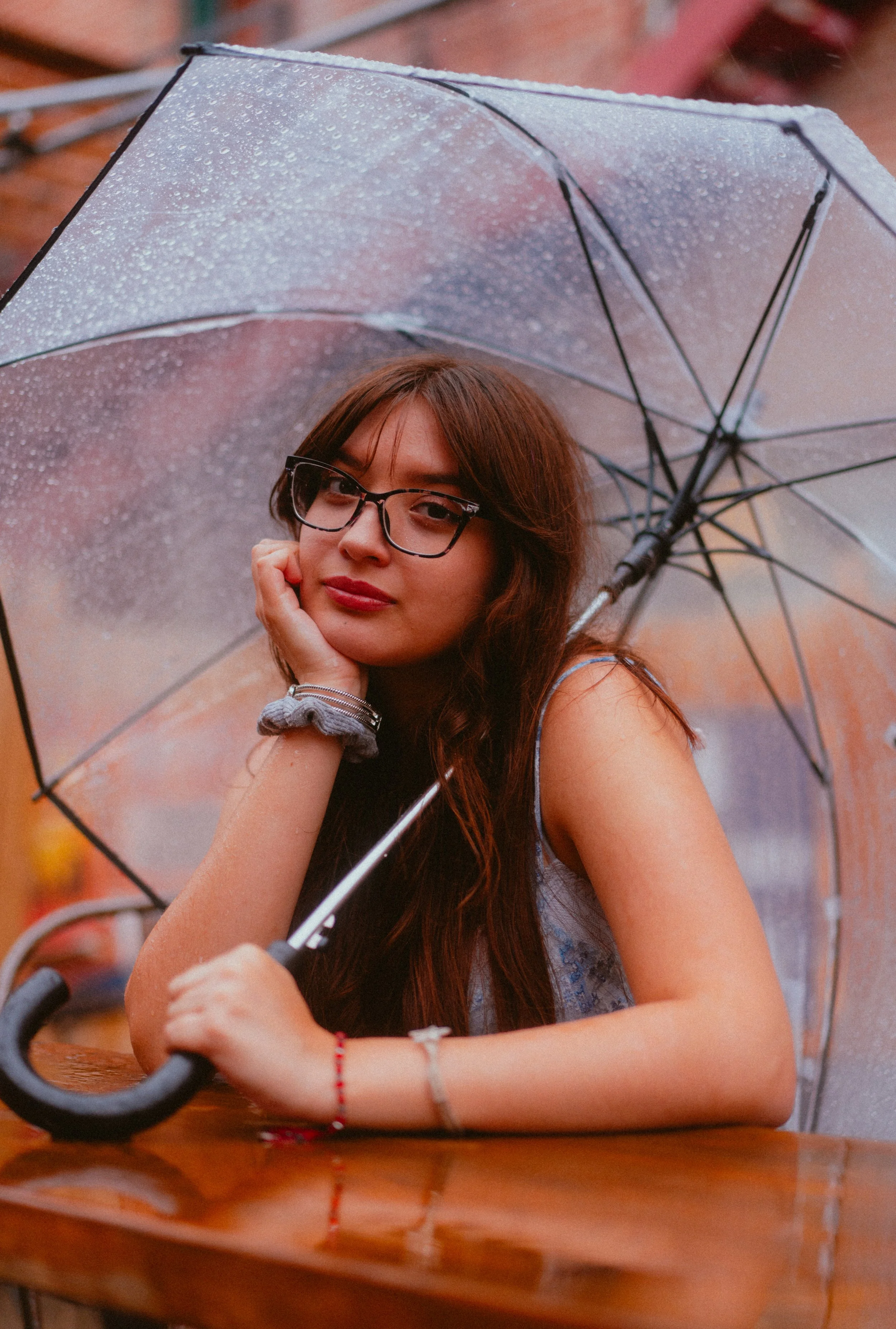 A young woman with long brown hair, glasses, and a red bracelet, sitting at a wooden table and holding an umbrella with raindrops on it.