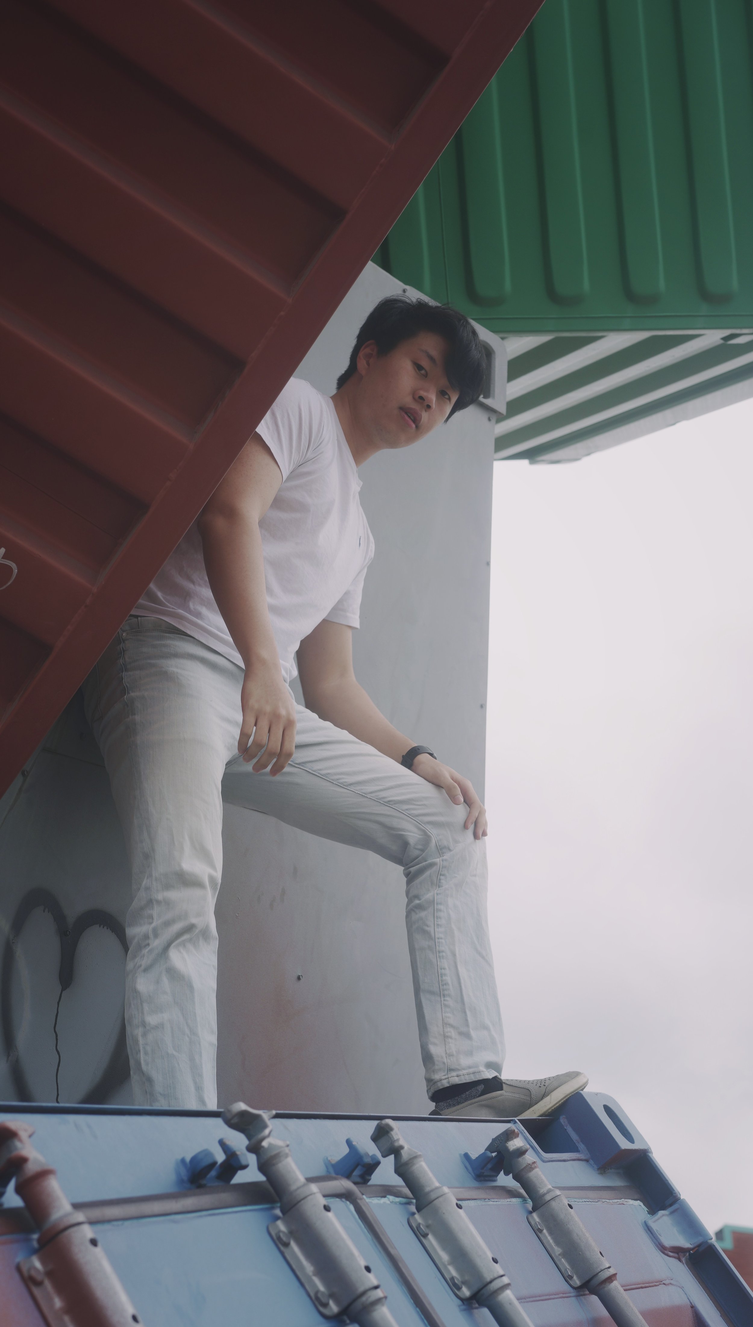 A young man with black hair, wearing a white T-shirt, beige pants, and sneakers, is sitting on a rooftop ledge against a gray wall, with a green awning above and a piece of industrial equipment with hoses below him. He is looking towards the camera.