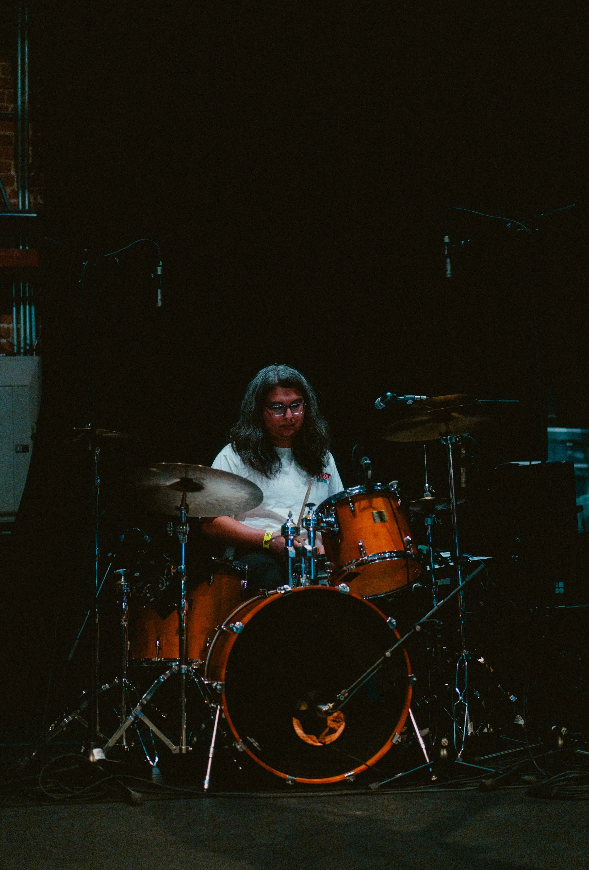 A woman with long dark hair and glasses playing a drum set on a dark stage, with microphones and equipment visible.