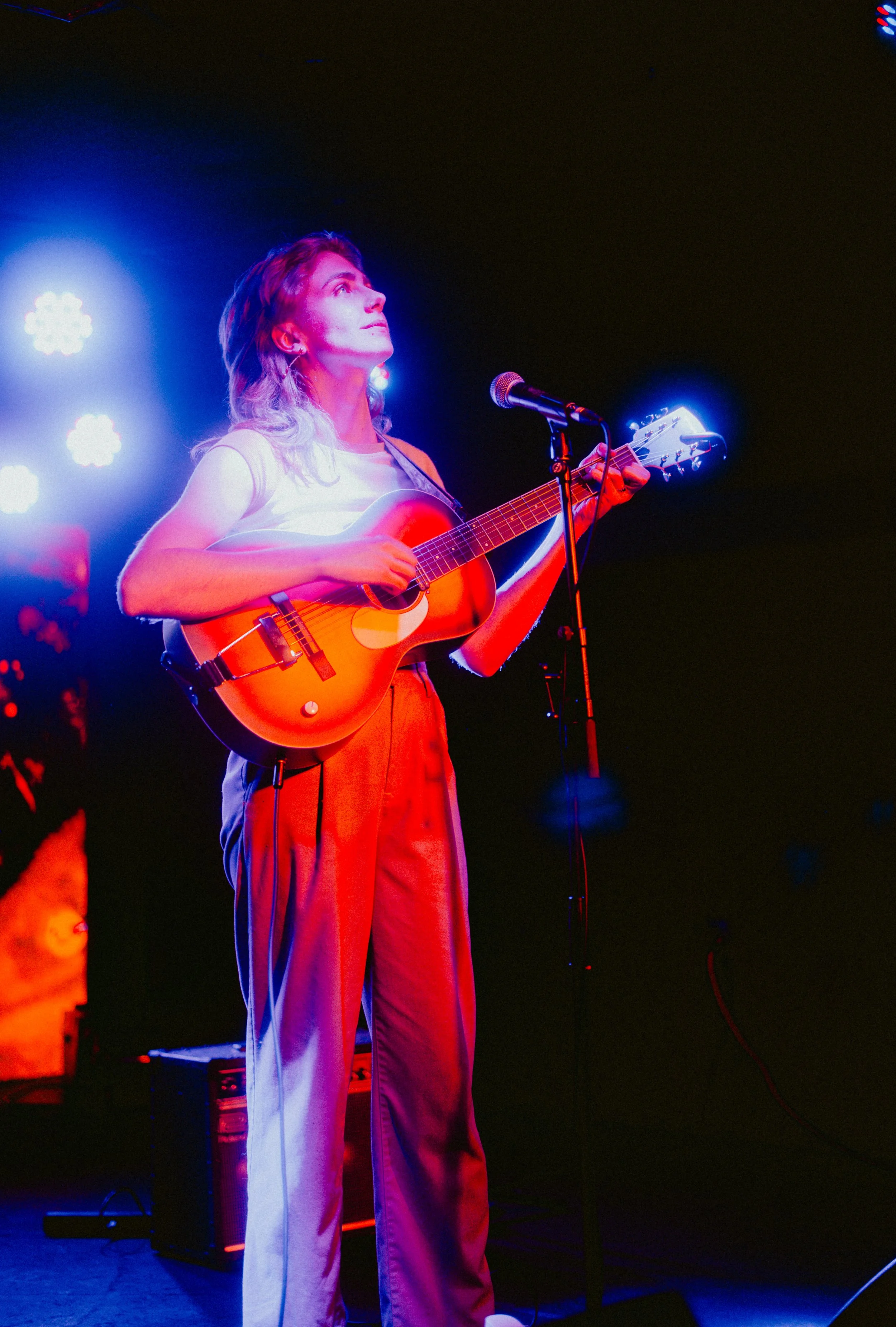 A woman with long hair playing an acoustic guitar on stage under colorful lighting, with a microphone in front of her and a small amplifier on the floor.