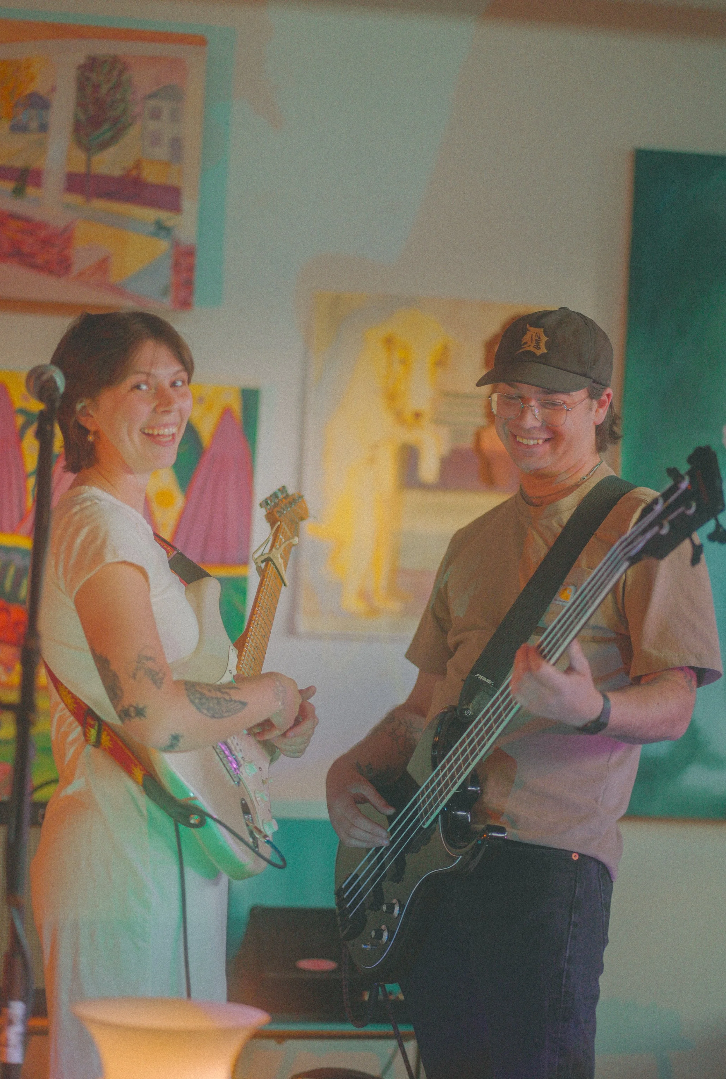 A woman with tattoos smiling and holding an electric guitar, and a man with glasses and a cap playing an acoustic guitar, standing in an art-filled room.