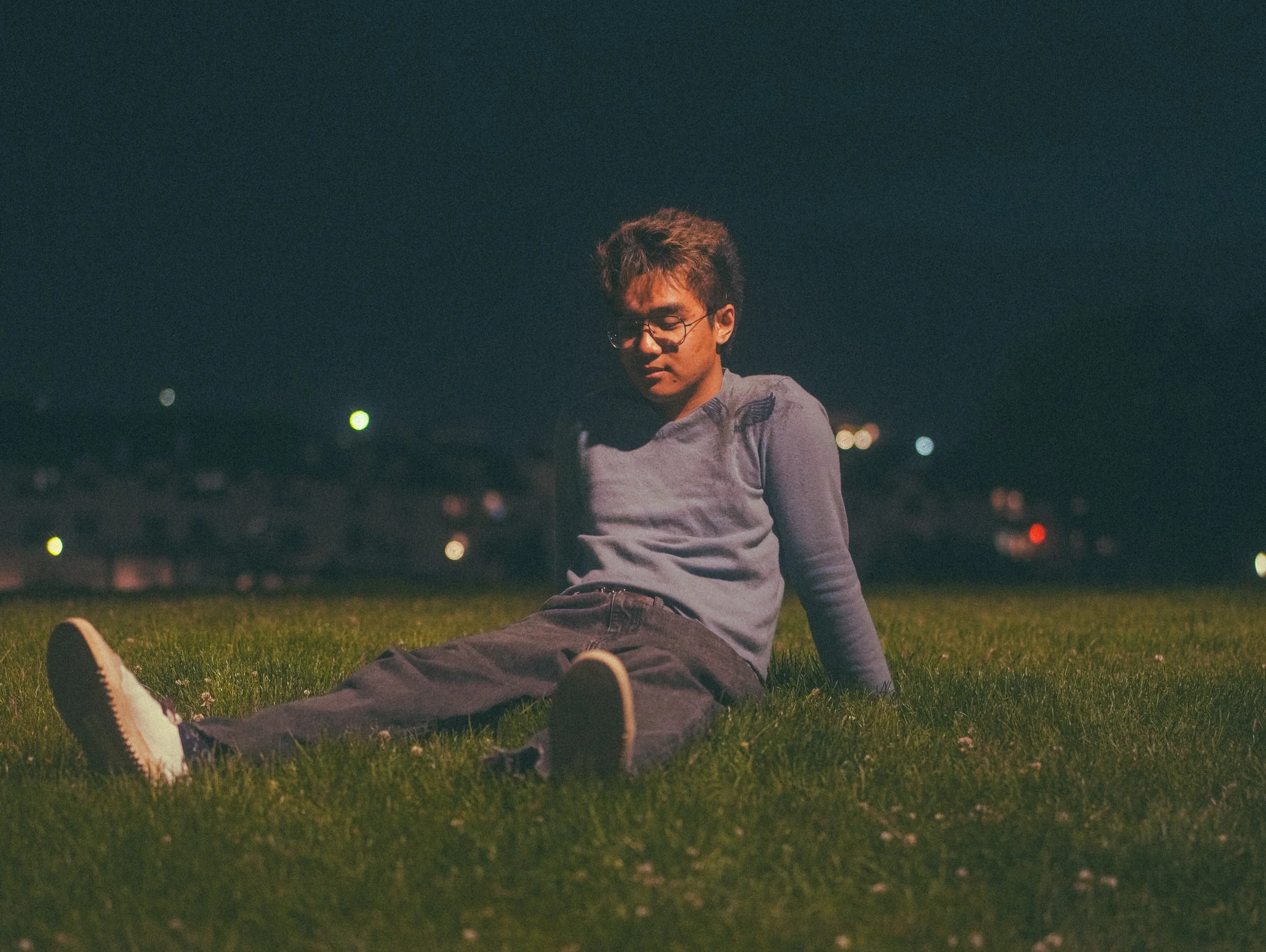 A young man with glasses and styled hair sitting on the grass at night, with city lights in the background.