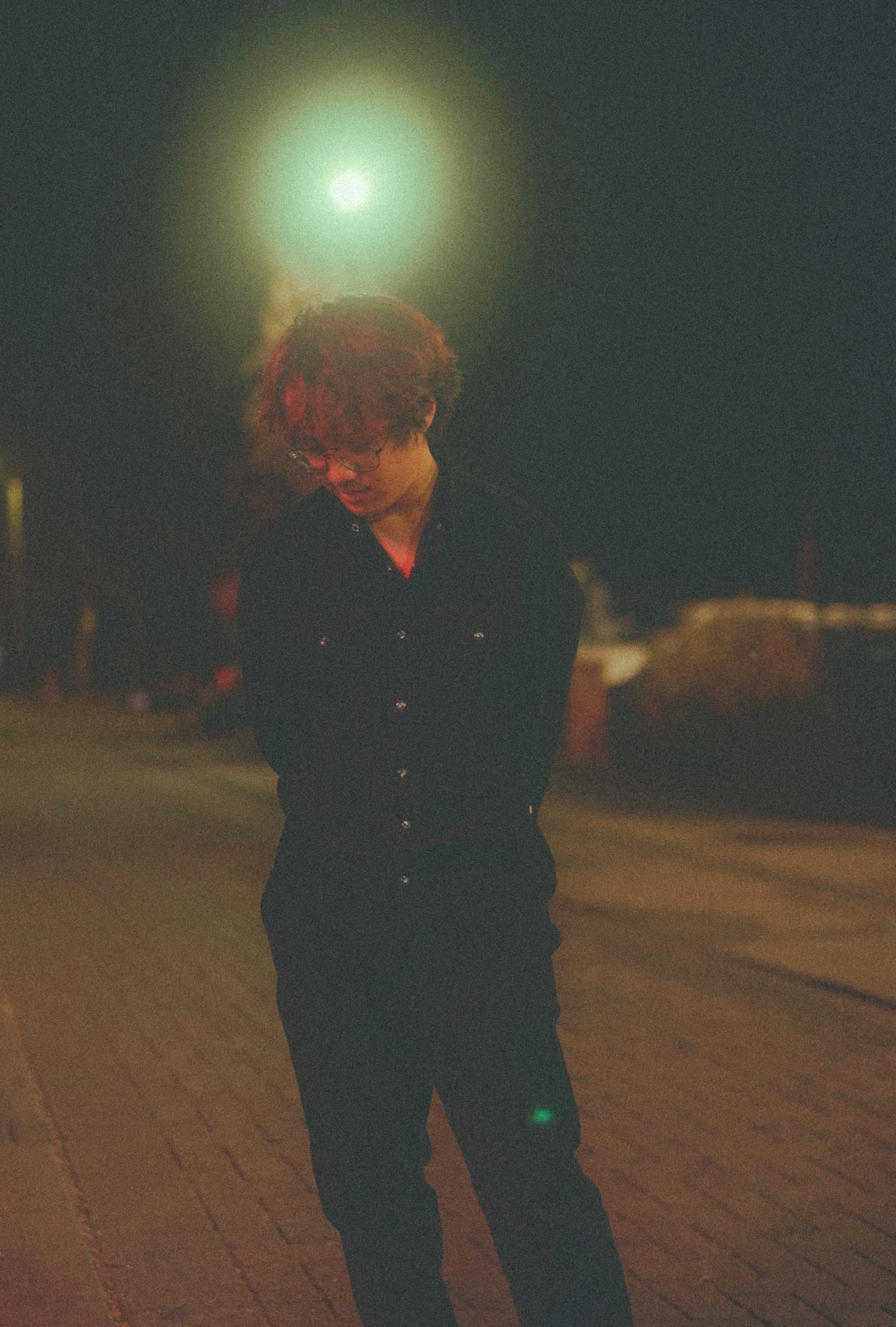 A person with reddish hair hairstyle and glasses stands looking downward in front of a bright moon in the dark night sky.