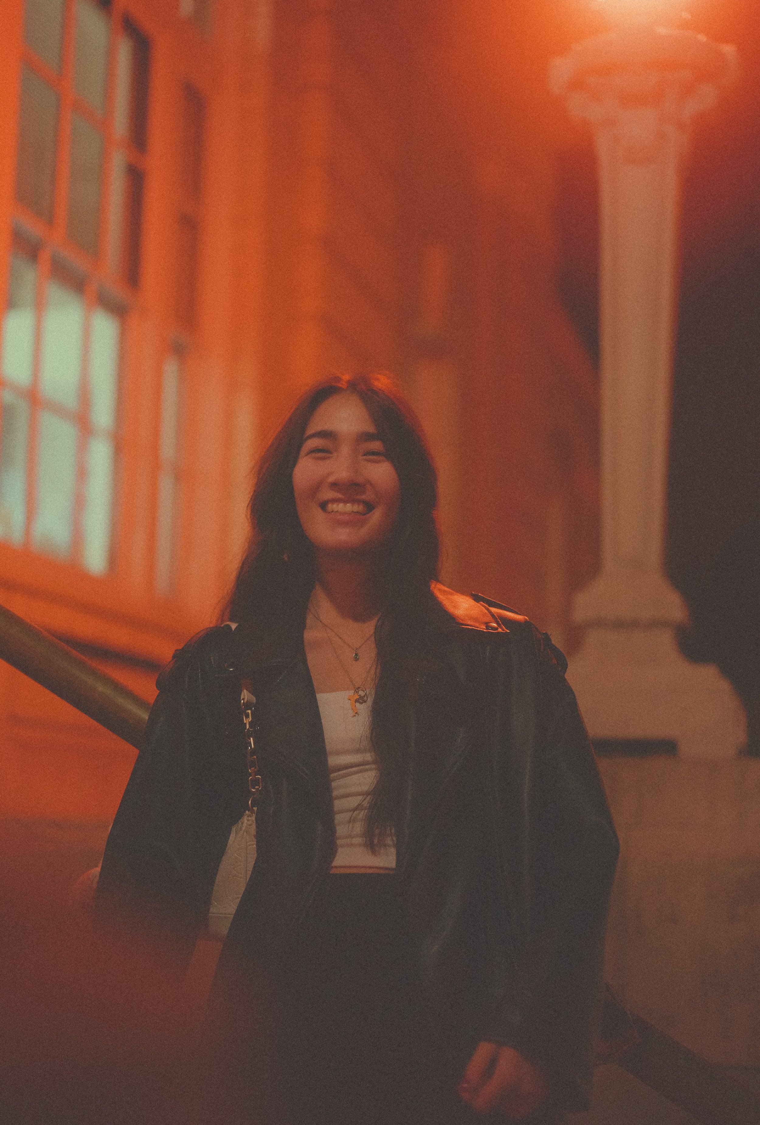 A young woman with long dark hair smiling, wearing a black leather jacket and a white top, standing in a brightly lit orange-walled space at night.