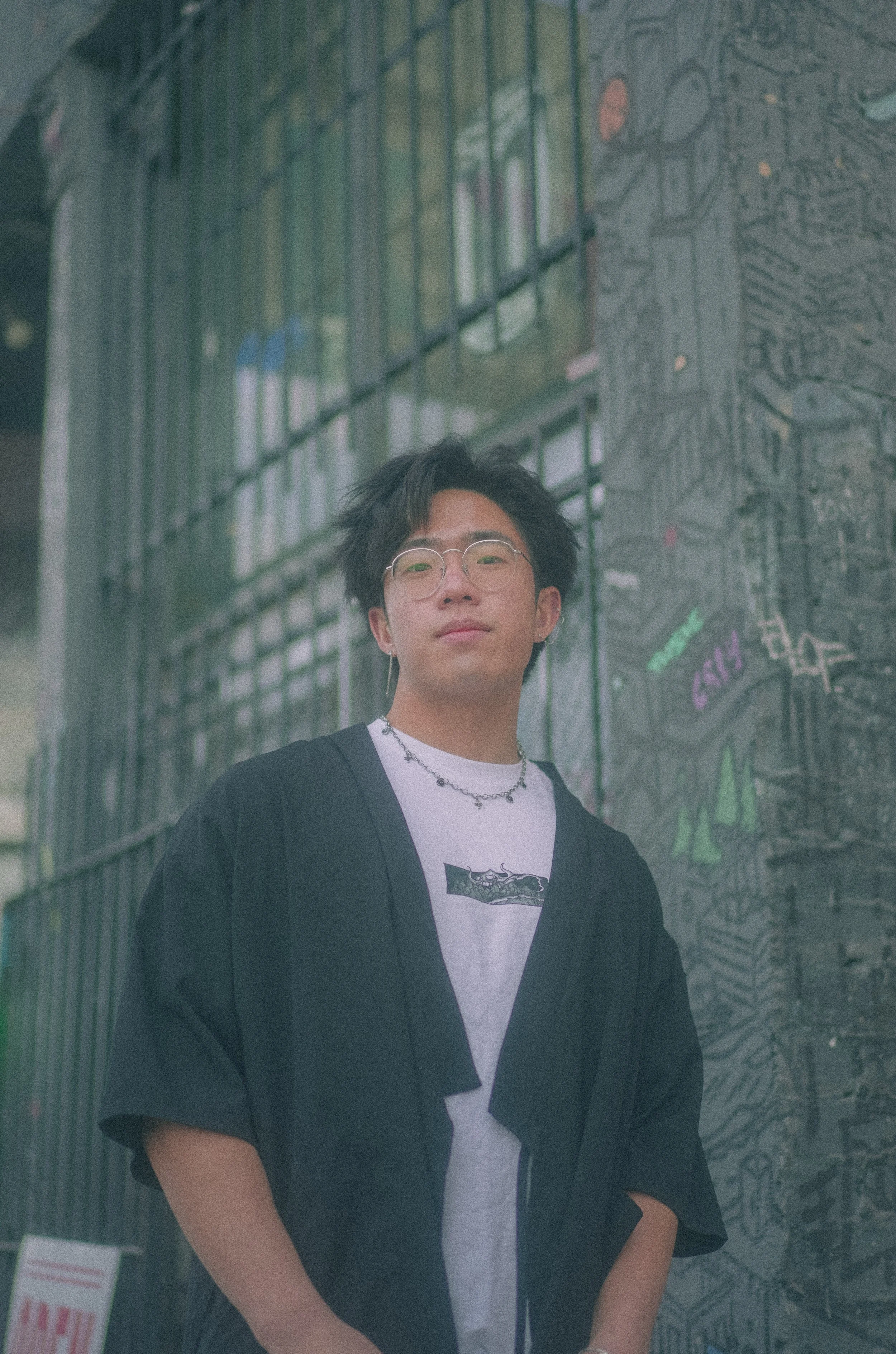 A young man with black hair, glasses, and a slight smile, standing outdoors in front of a mural or graffiti wall.