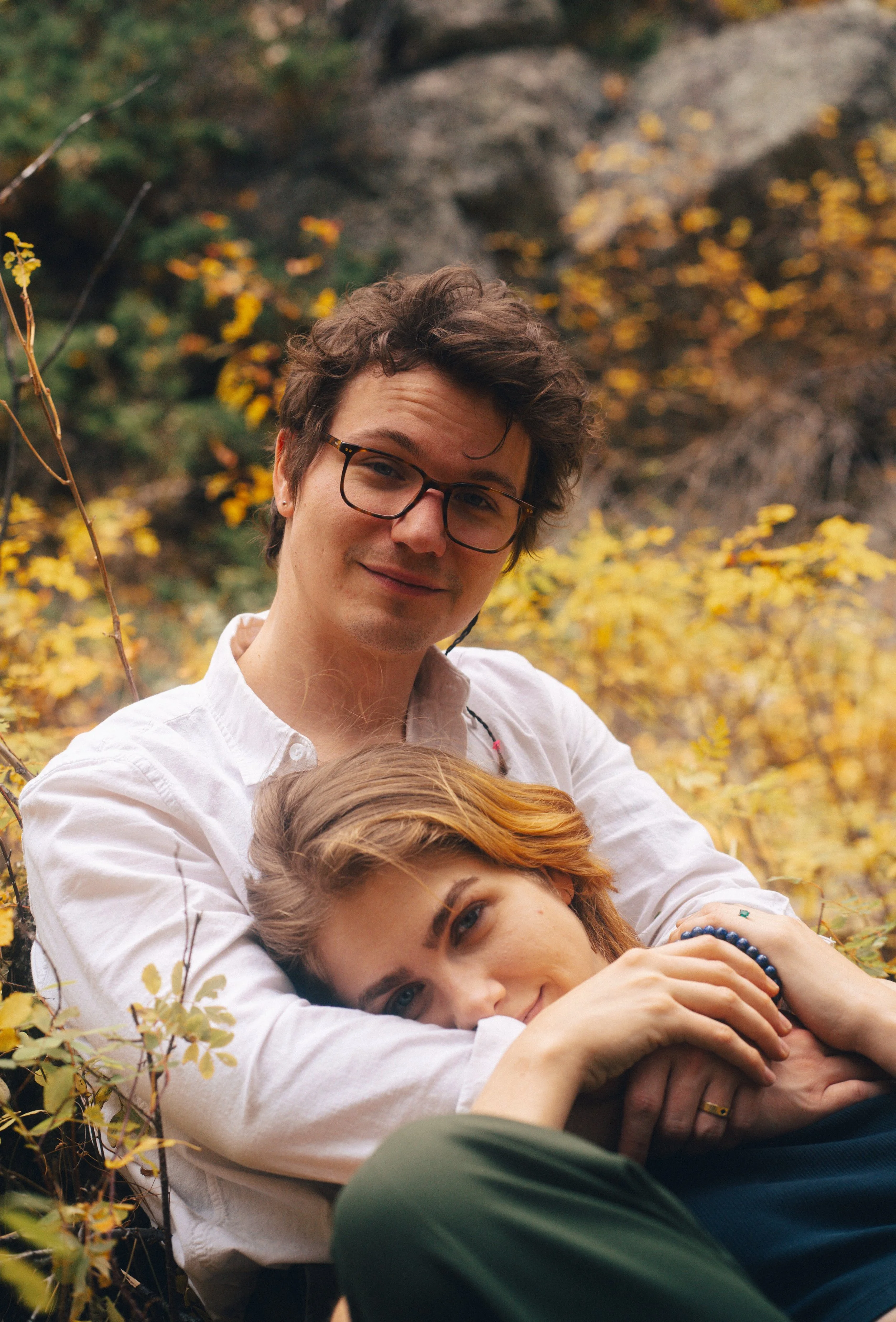 A young man with glasses and two young women lying outdoors among yellow autumn leaves, cuddling and smiling at the camera.
