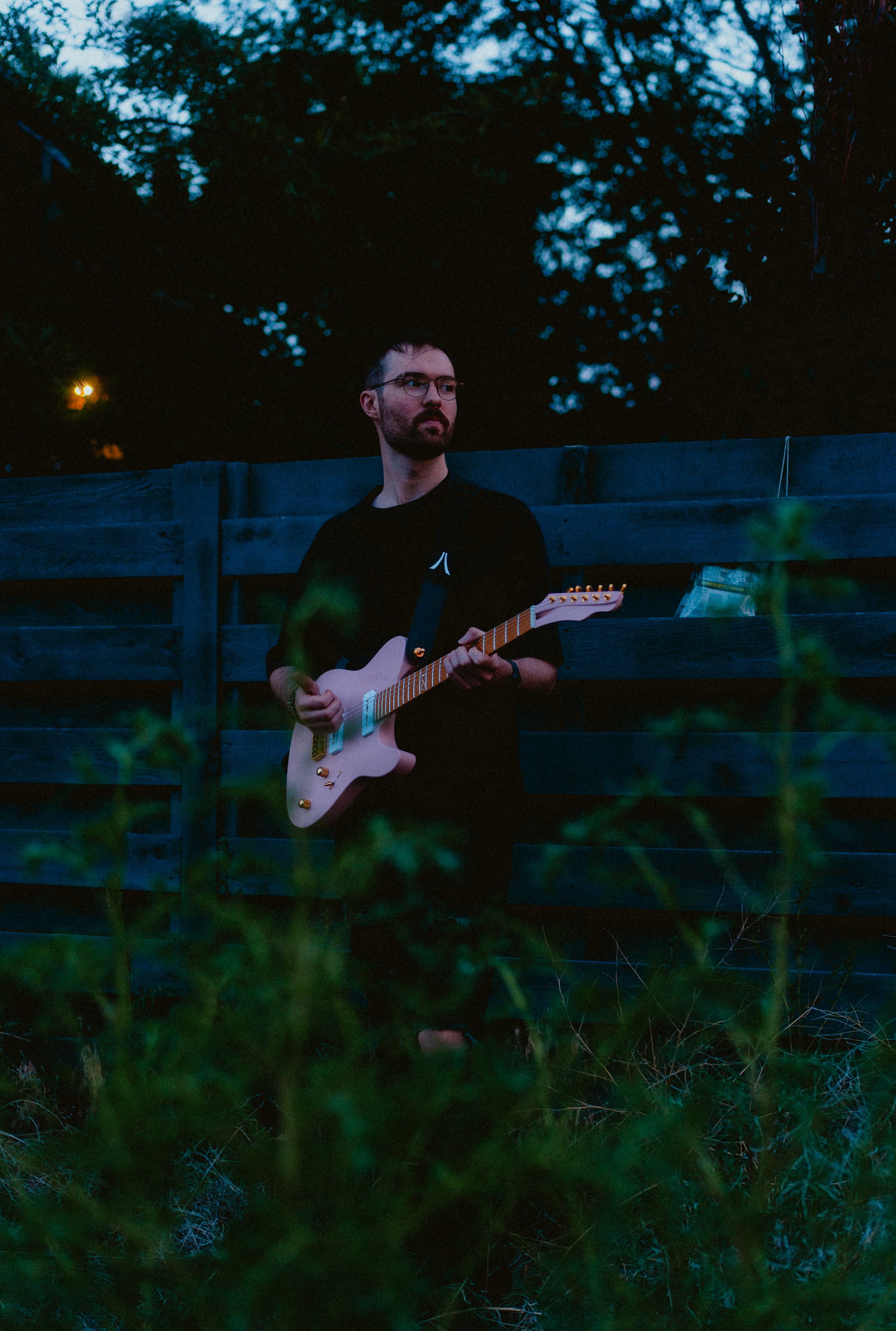A man with glasses and a beard playing an electric guitar outdoors at dusk, standing in front of a wooden fence with blurry greenery in the foreground and trees in the background.
