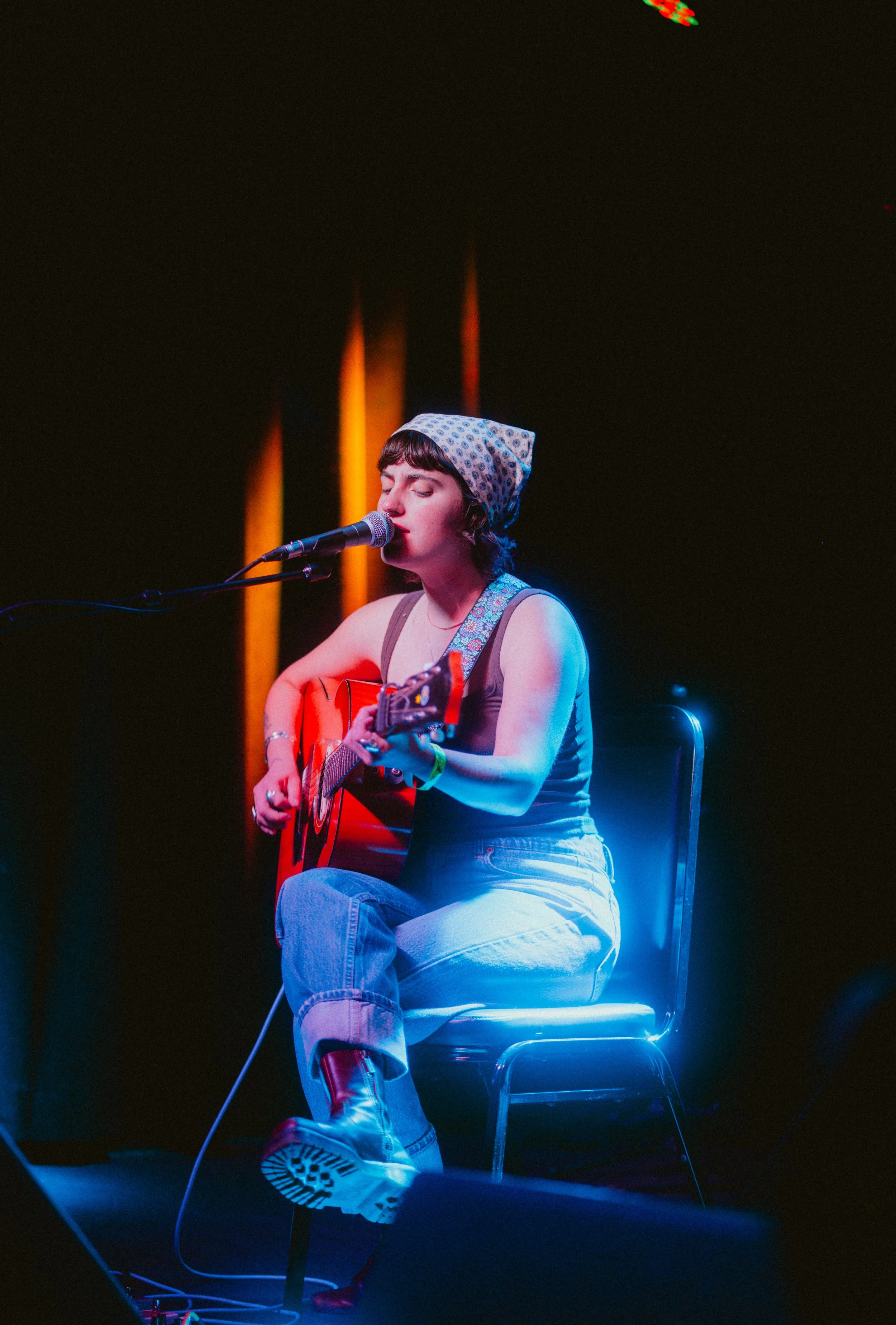 A young woman with short brown hair, wearing a patterned headscarf, is sitting and singing into a microphone while playing an acoustic guitar. She is dressed in a tank top, jeans, and sneakers, in a dimly lit setting with dark background and colorful