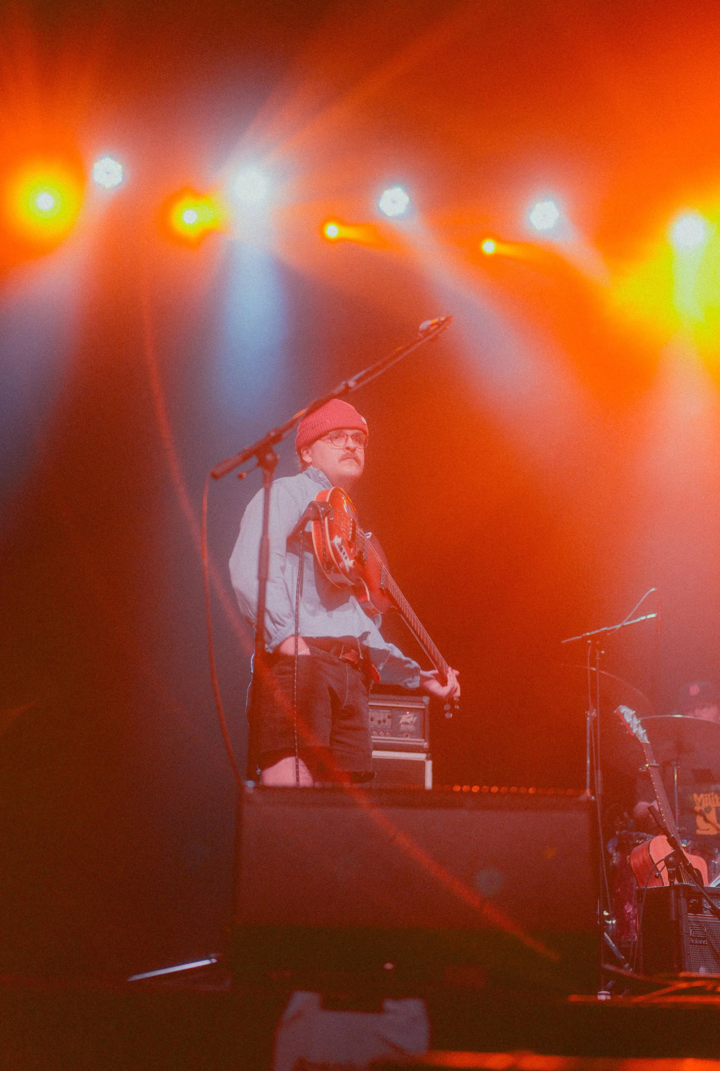 A musician wearing a red beanie, glasses, and a jacket is on stage with a guitar, under orange stage lighting, with musical equipment around him.