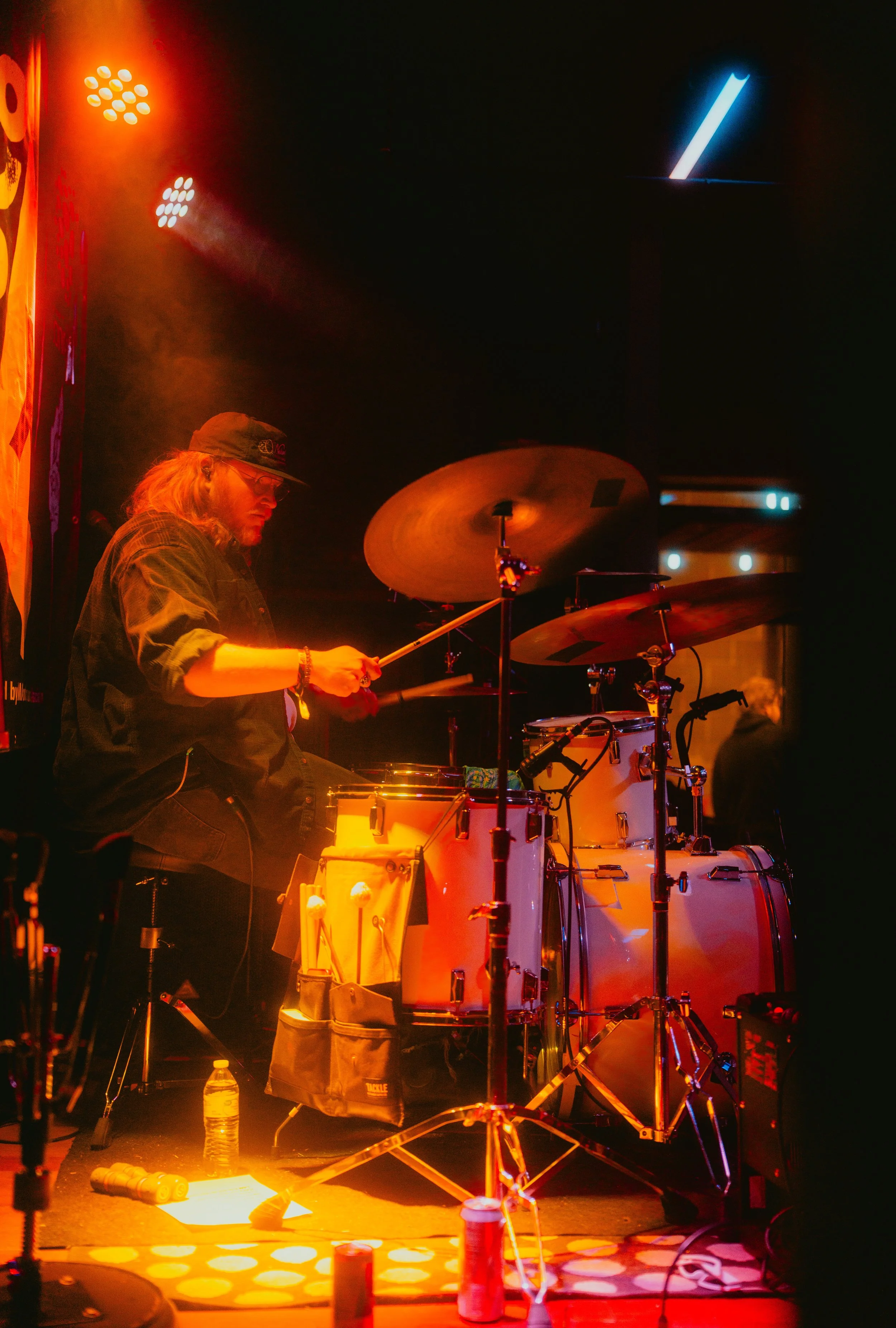 A musician playing drums on stage with stage lighting and equipment around.