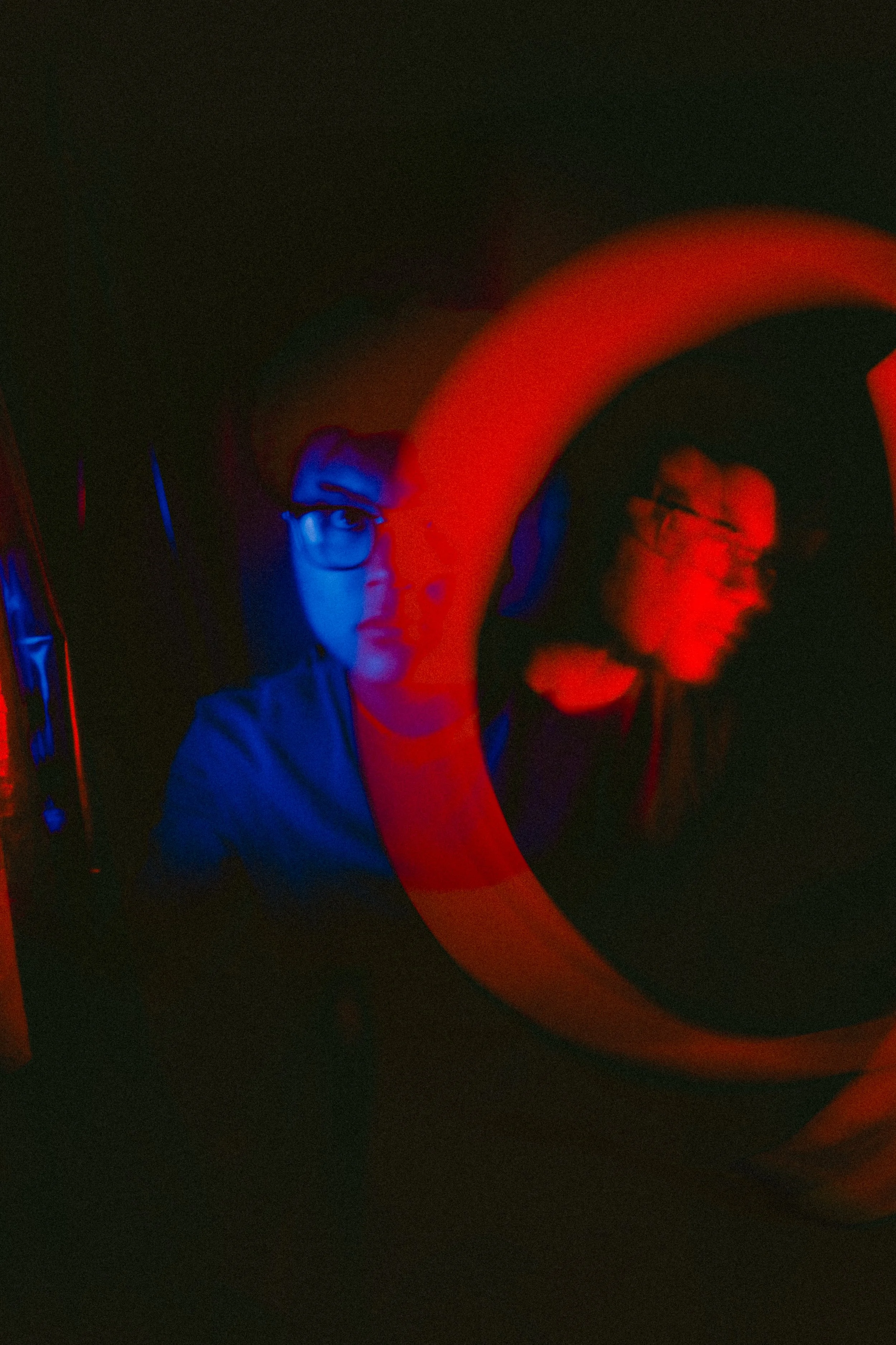 A person with glasses, wearing a blue shirt, is illuminated with red and blue lights in a dark room. There is a circular red neon light around their face, creating a glowing effect.