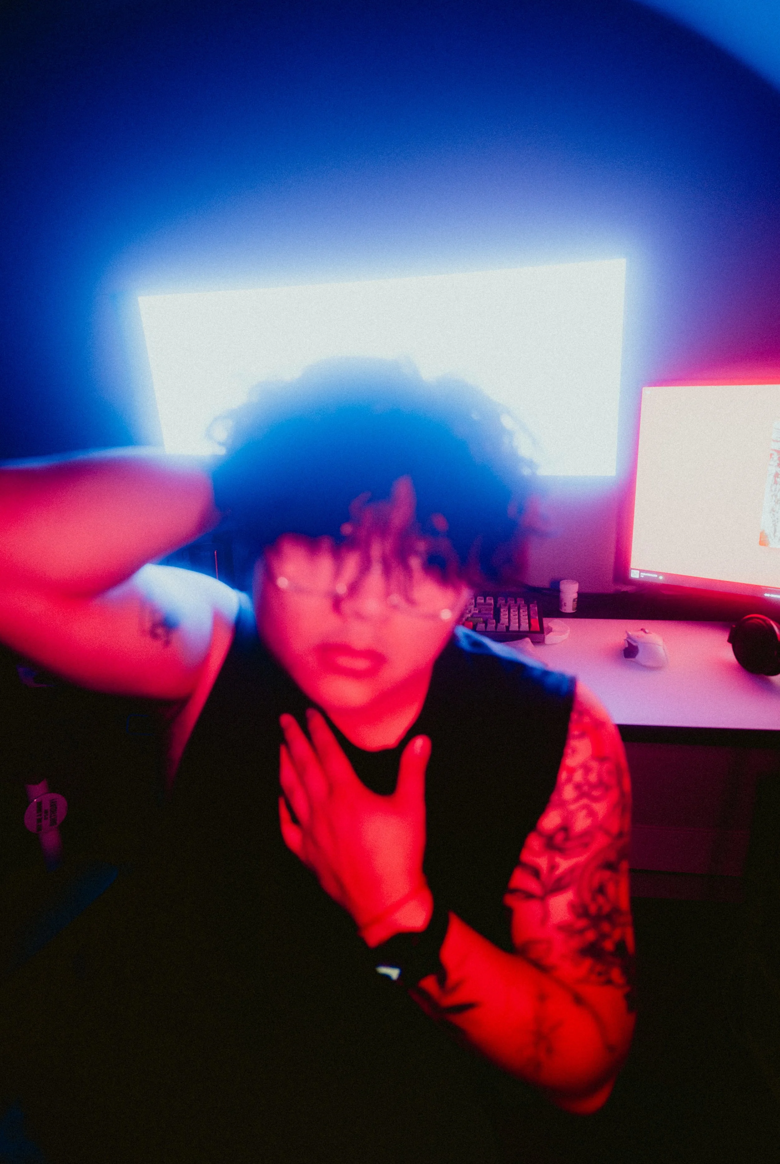 Person with curly hair wearing a dark sleeveless shirt, sitting in front of multiple computer screens with bright displays, in a dark room illuminated by screen light, making a peace sign with their fingers.