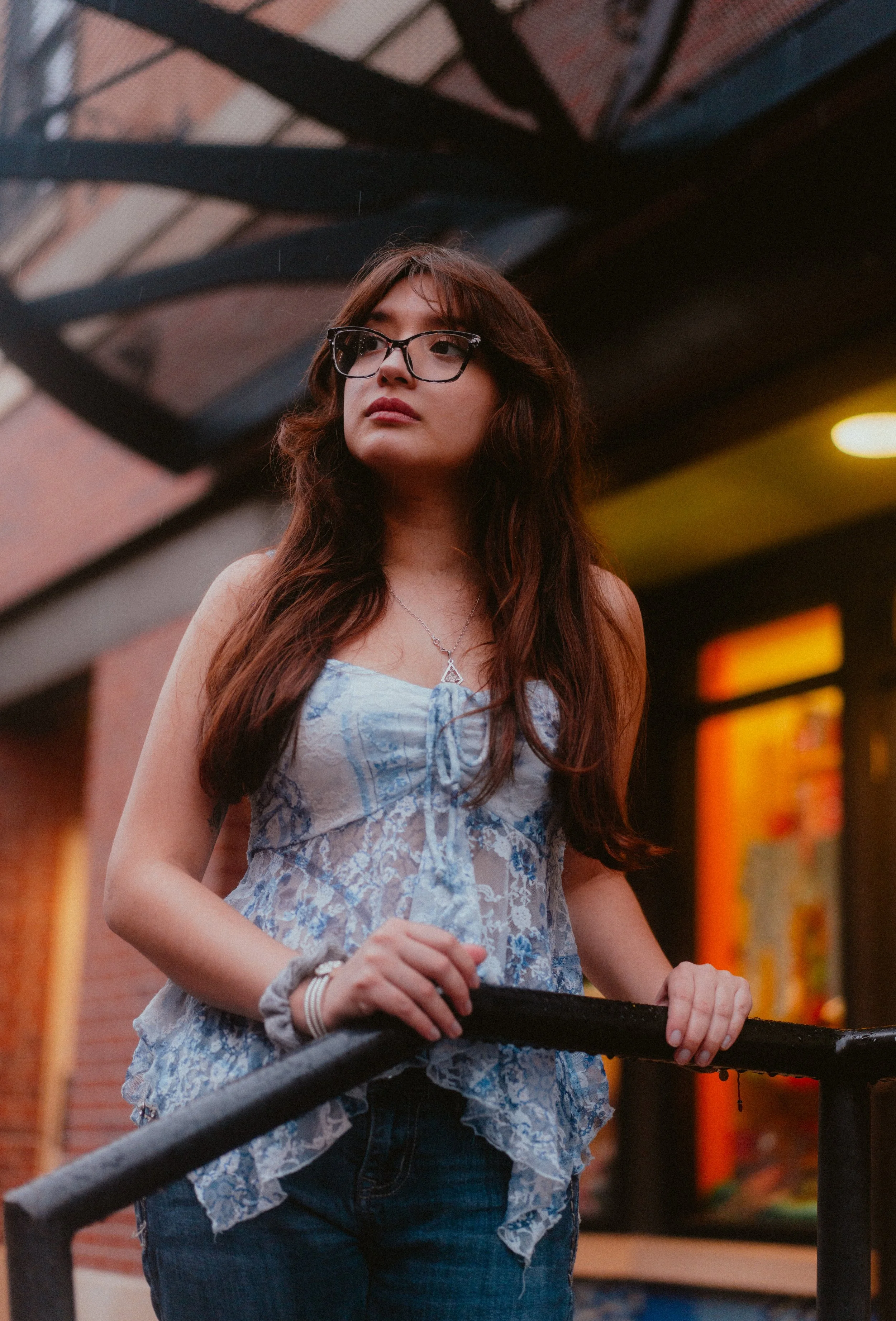 A young woman with long brown hair and glasses looking to the side on a city street at dusk, wearing a light blue lace top and jeans, holding onto a black railing.