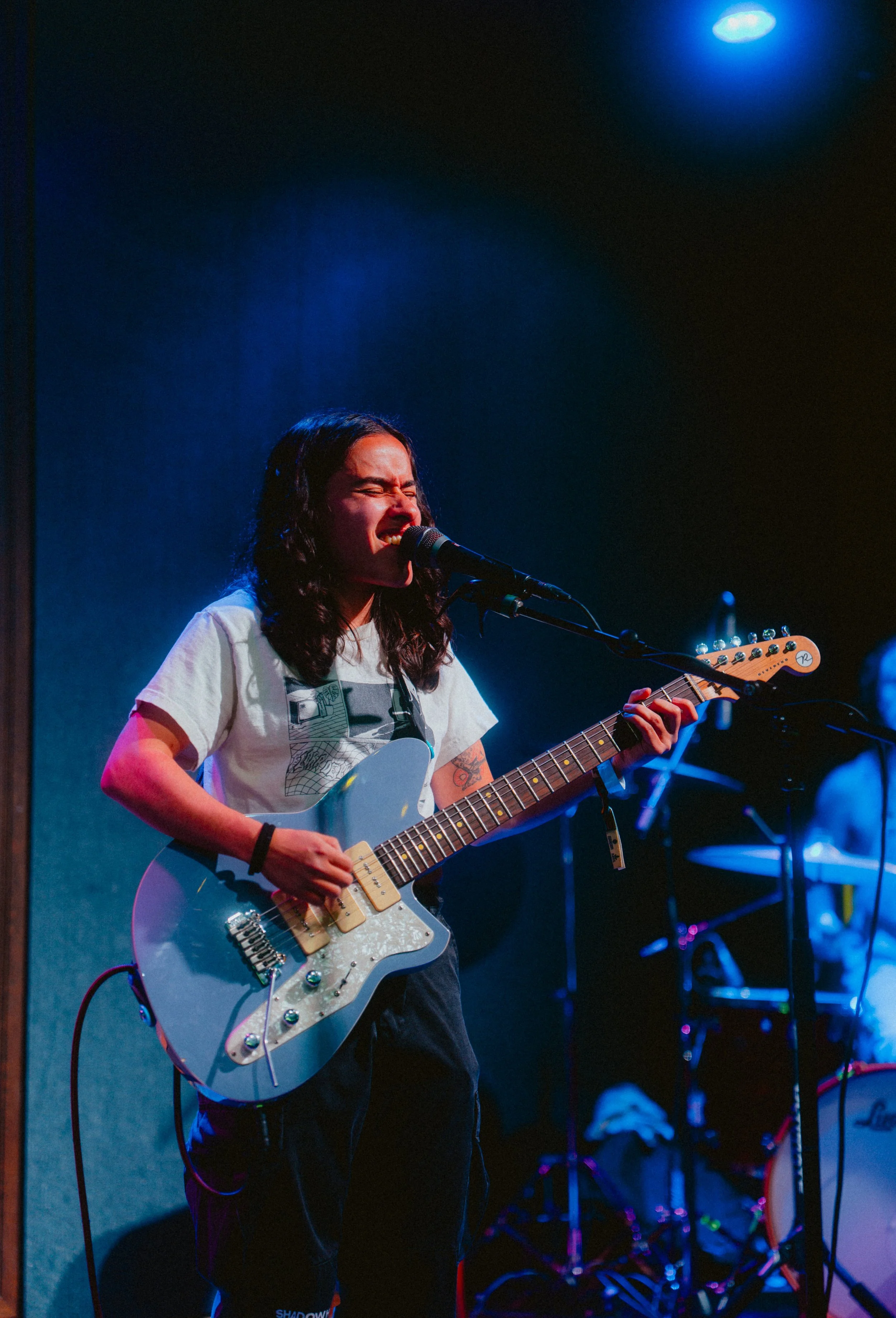 A young woman singing into a microphone while playing an electric guitar on stage with blue and purple lighting.