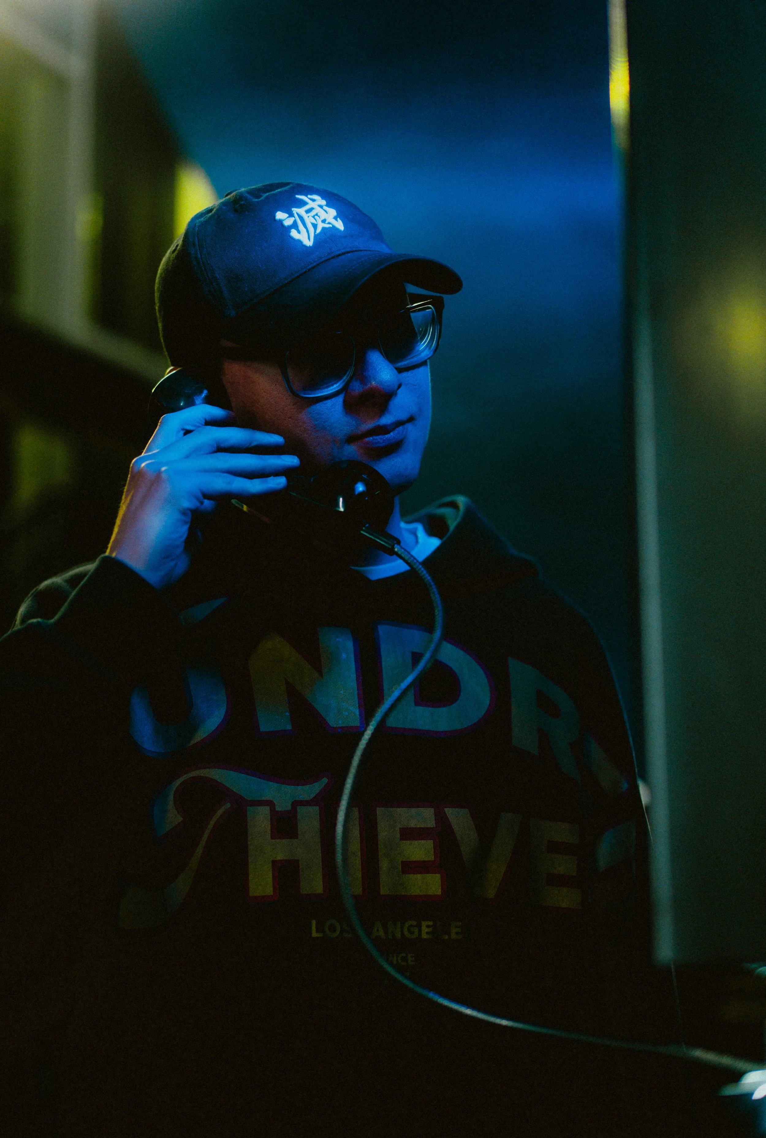 A young person wearing a dark cap with white Asian characters, large glasses, and a hoodie, talking on a phone in a dimly lit environment with blues and dark colors.