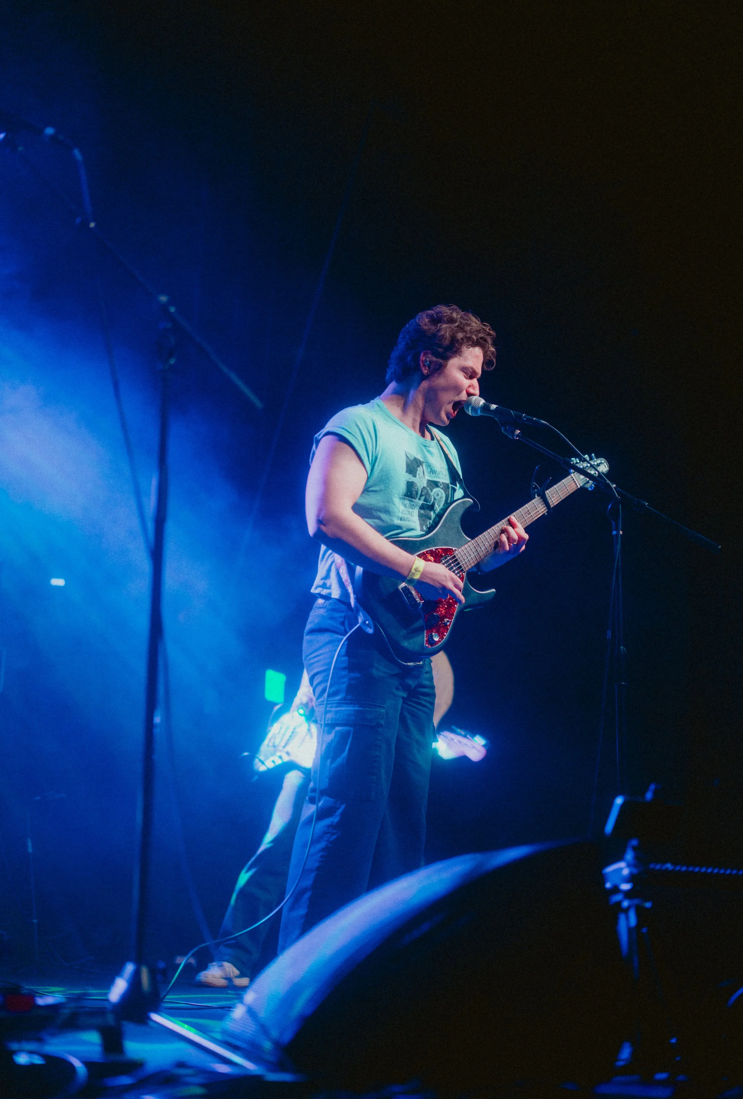 A woman with short curly hair wearing a light blue t-shirt playing an electric guitar and singing into a microphone on stage with blue and purple lighting.