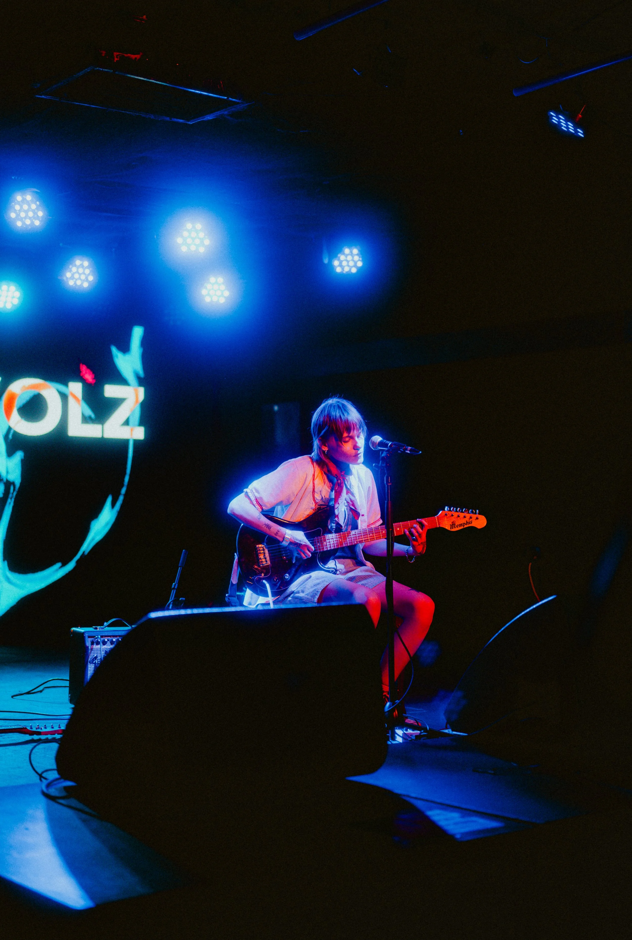 A female musician sitting on a chair playing an electric guitar on a dimly lit stage with blue lighting, under a sign that partially reads 'OLZ' and a partial logo visible.