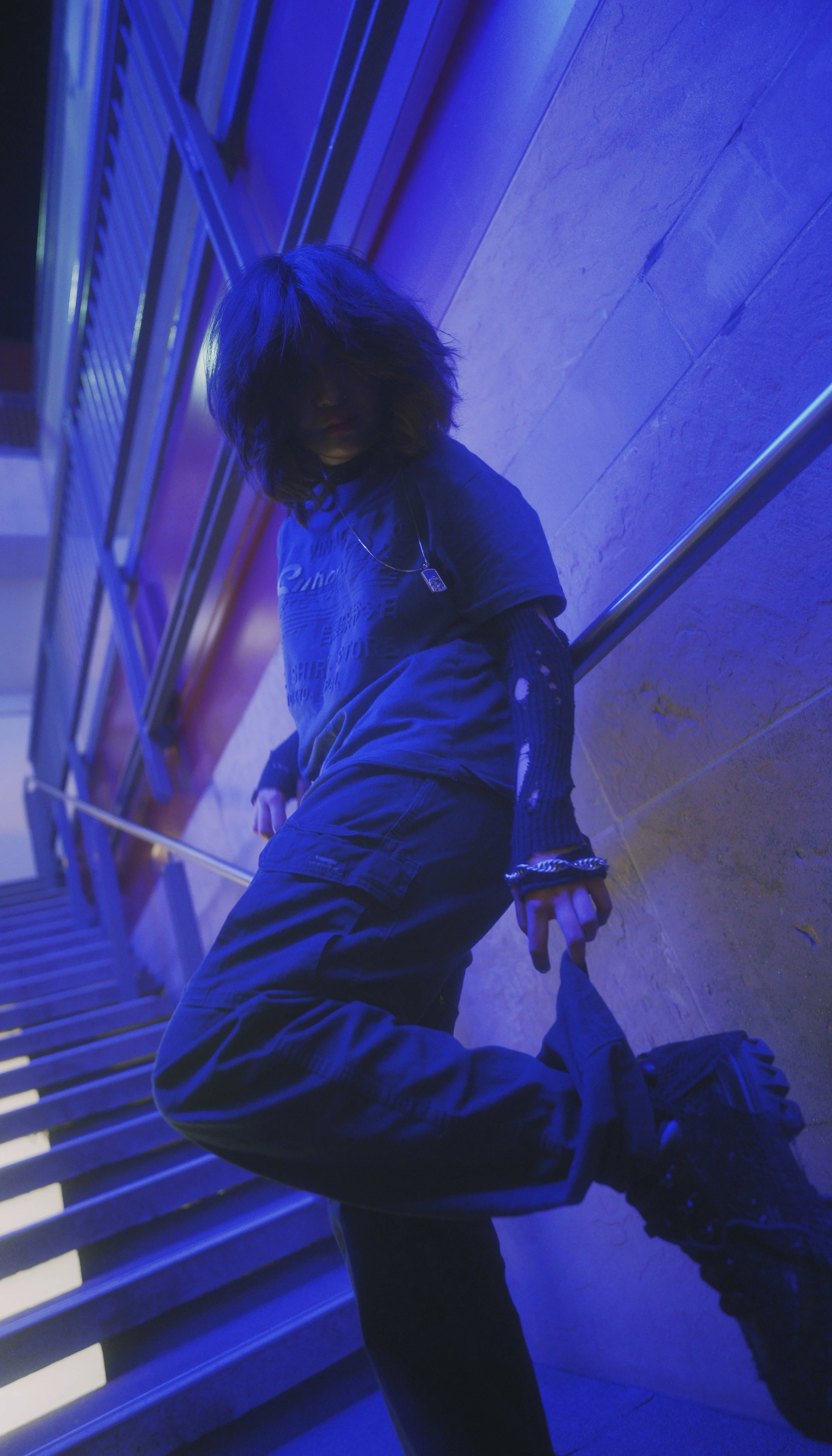 A person with shaggy hair styled to cover their face, dressed in dark clothing and a necklace, leaning against a wall illuminated by blue and purple lighting. They are sitting on the stairs and holding an object in their hand.