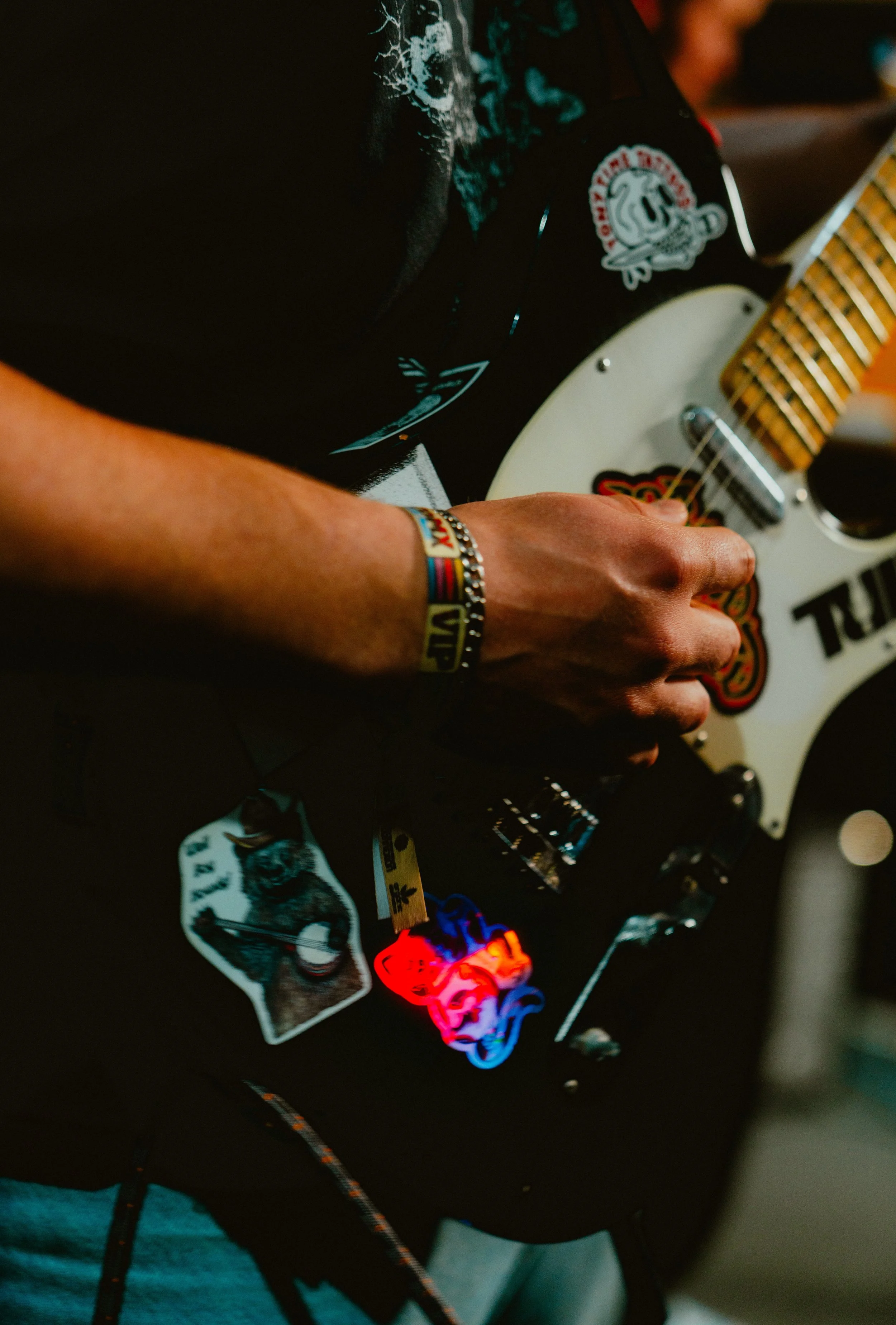 Close-up of a person playing an electric guitar with various stickers, including a skull and a dog, on the guitar's body. The person wears a wristband and a bracelet.