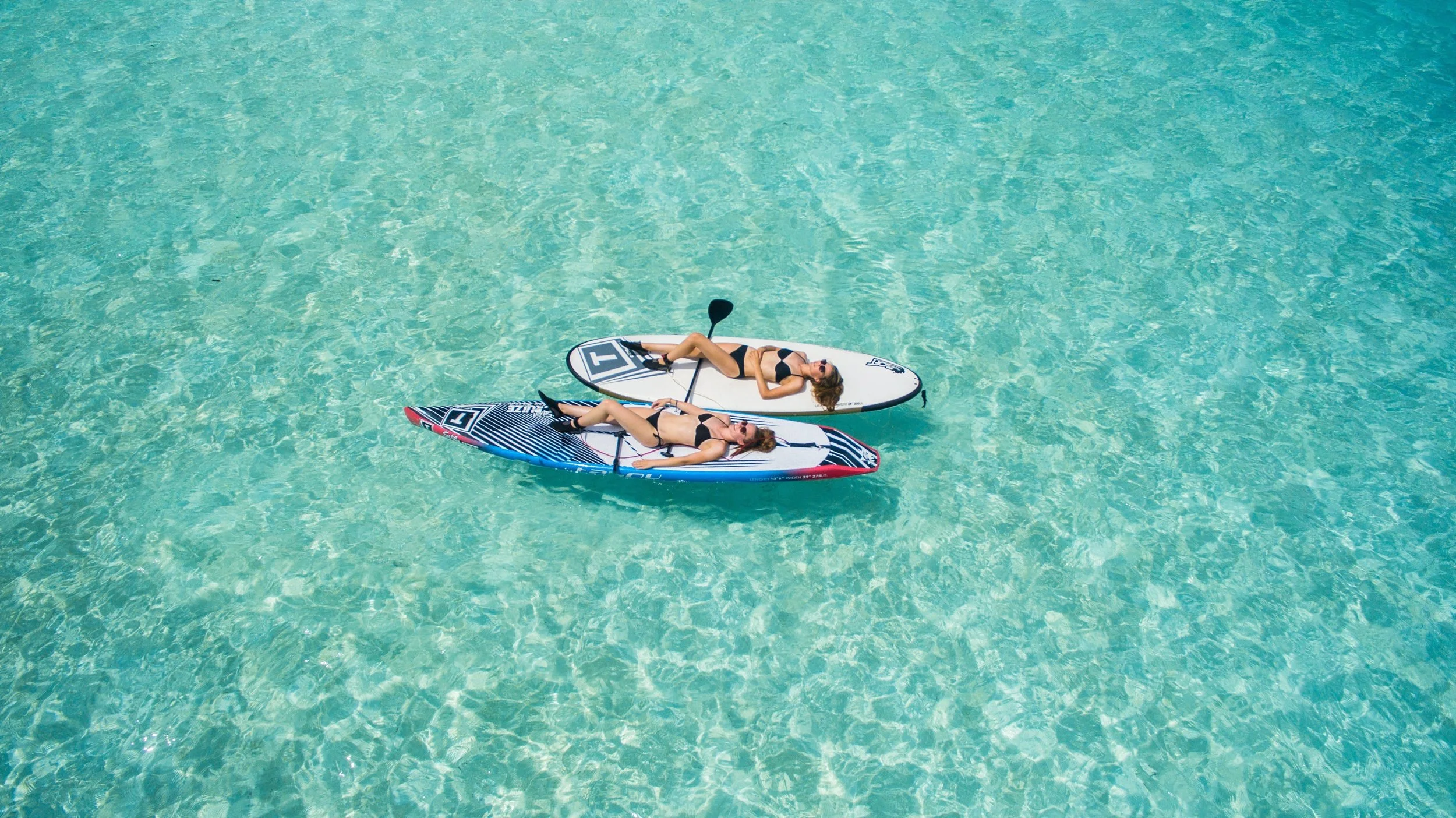 Two women in bikinis lounging on stand-up paddleboards in clear turquoise water.