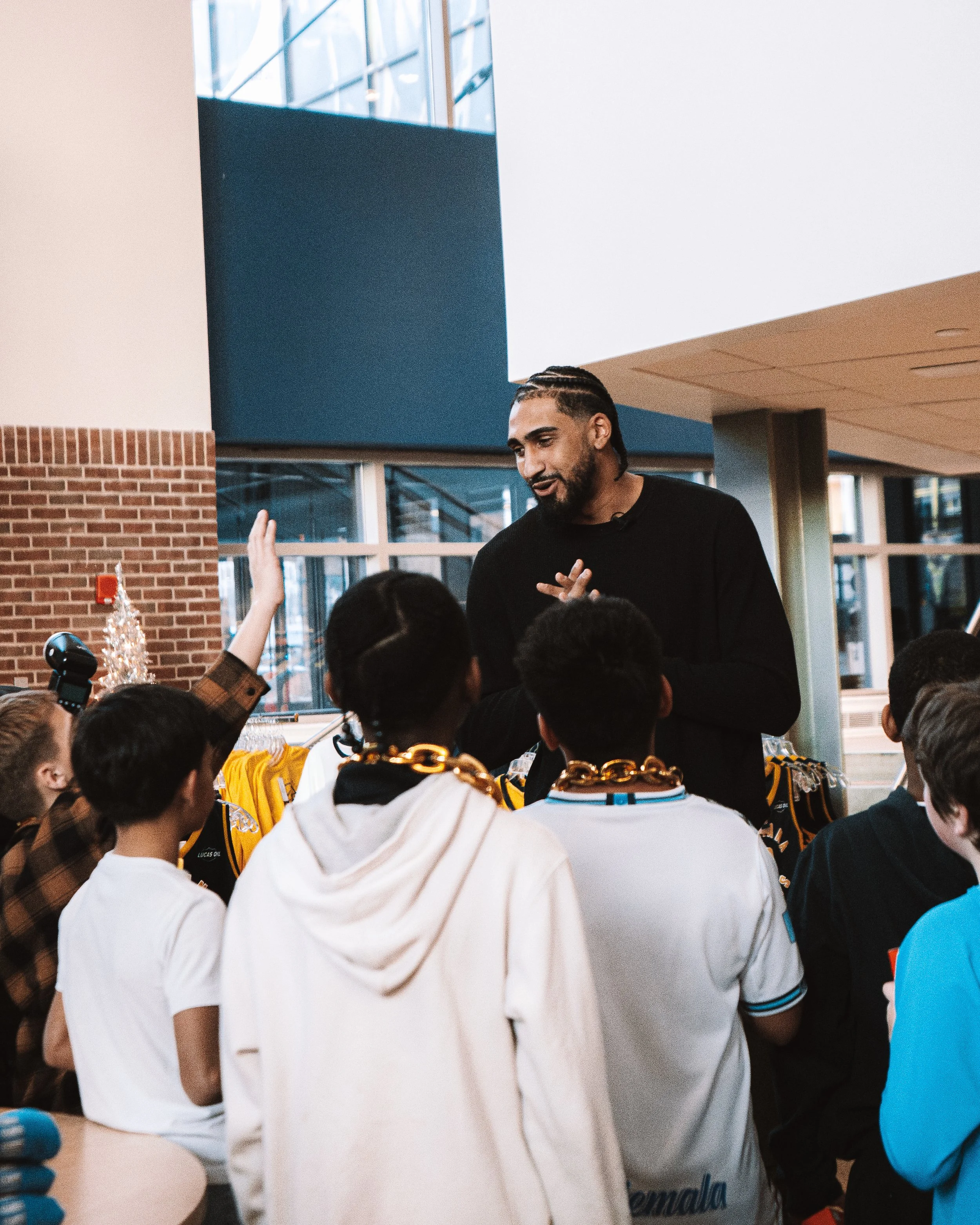 A man with braided hair and a beard is speaking to a group of children, some of whom have their hands raised, inside a building with large windows and brick walls.