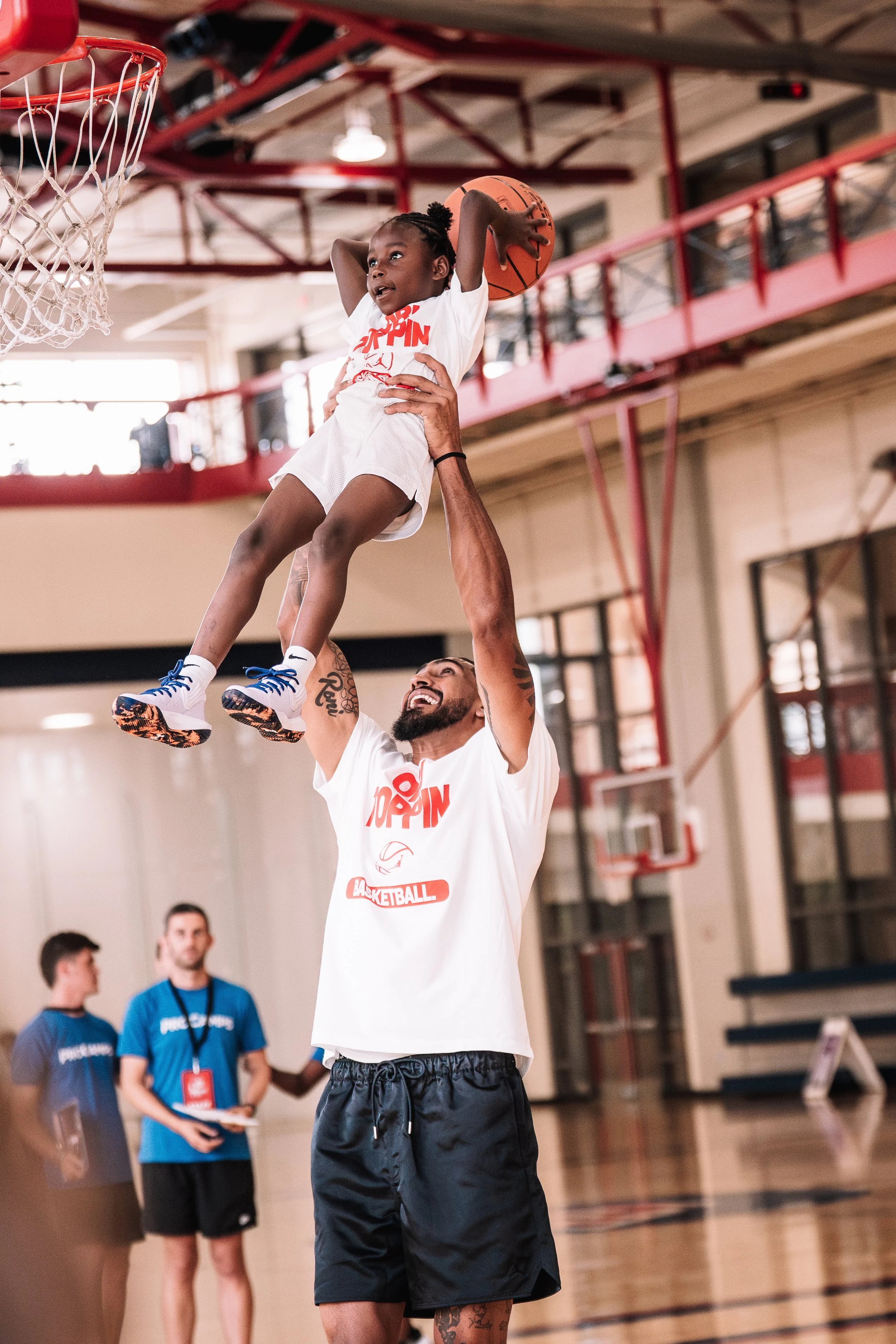 Young boy in a white basketball jersey and shorts holding a basketball above his head inside a gymnasium, with a man supporting him from below, and other people in the background.