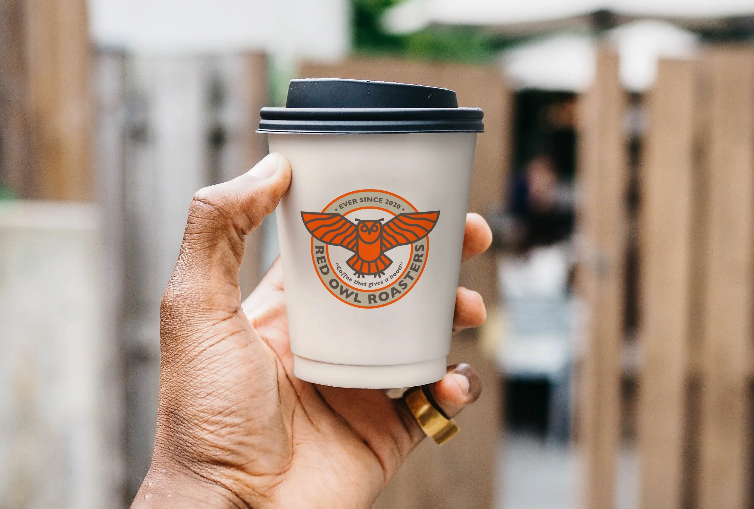 A hand holding a white coffee cup with a black lid, featuring a logo of a moth with orange wings, and the text "Ever Since 2020", "Red Owl Roasters", and slogan "Coffee that gives a hoot" on the cup.