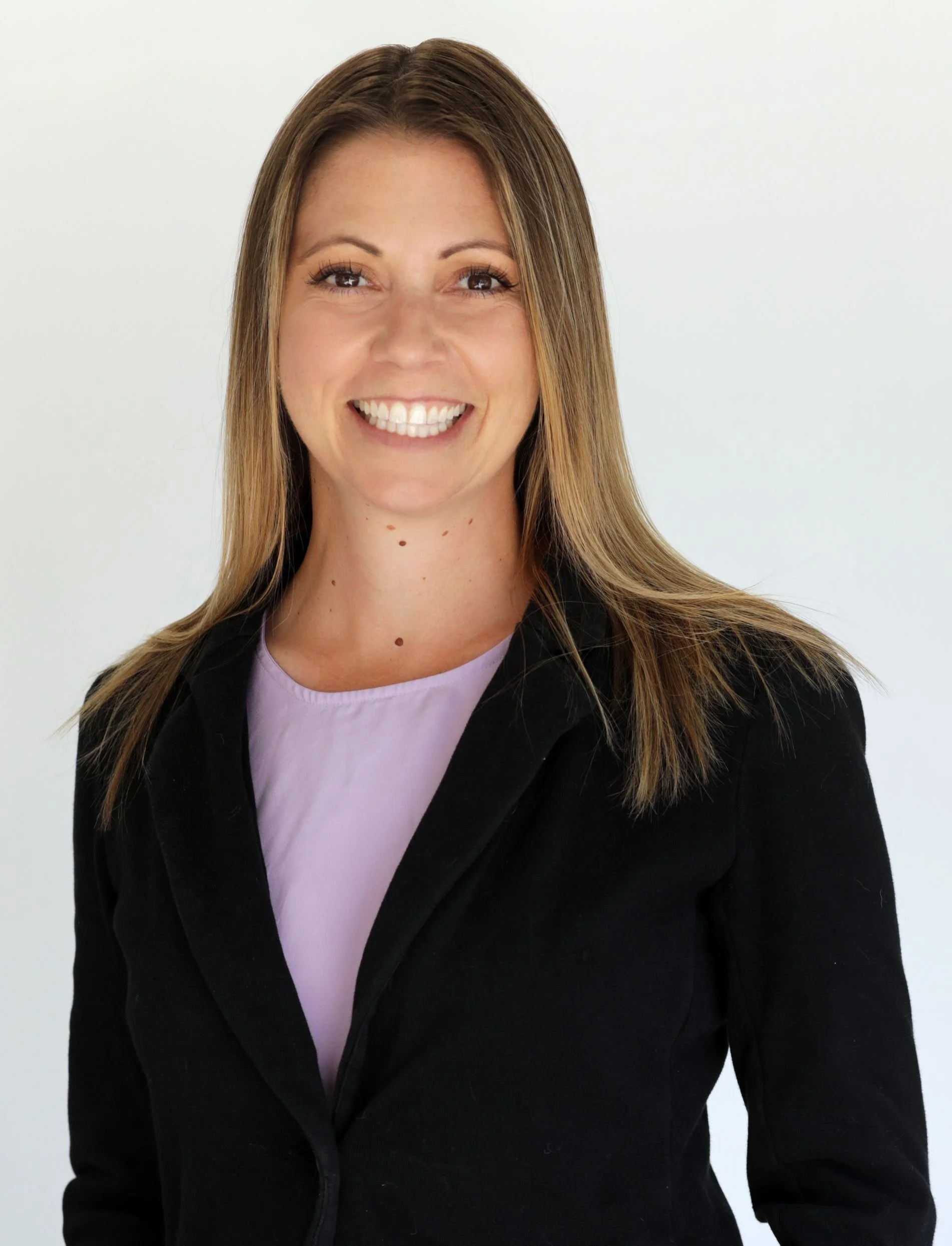 A woman with long brown hair, smiling, wearing a black blazer over a light purple shirt, standing against a plain white background.