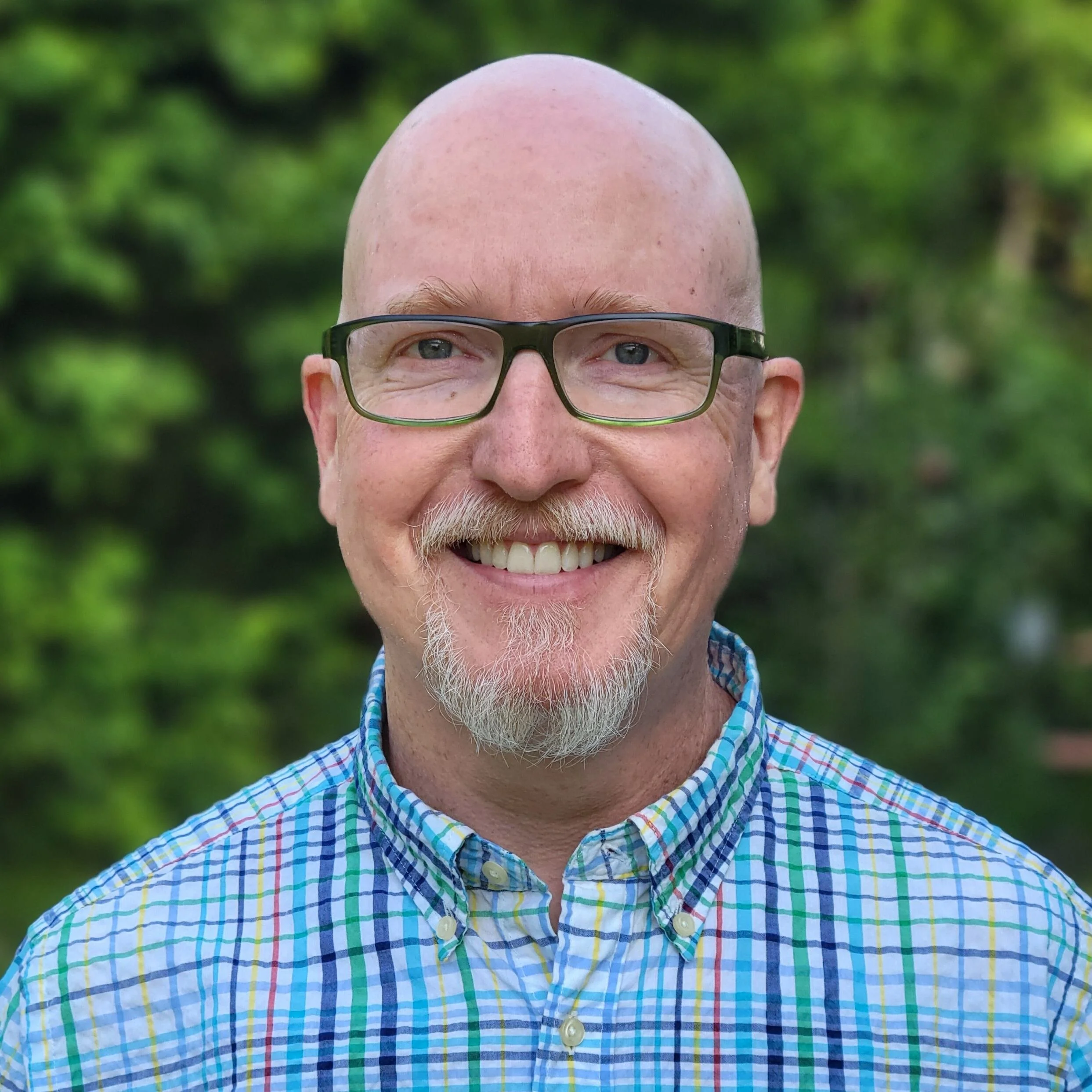 Close-up of a smiling middle-aged man with a bald head, glasses, and a white beard, wearing a blue, green, yellow, and red plaid shirt, outdoors with greenery in the background.