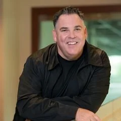 A smiling man with short, dark hair wearing a black jacket, indoors with a water feature in the background.