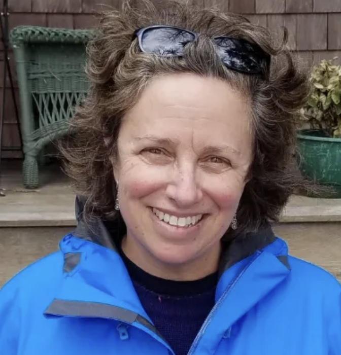A smiling woman with curly brown hair and sunglasses on her head, wearing a blue jacket, standing outdoors near a wooden deck with a green wicker chair and a large green pot with plants.