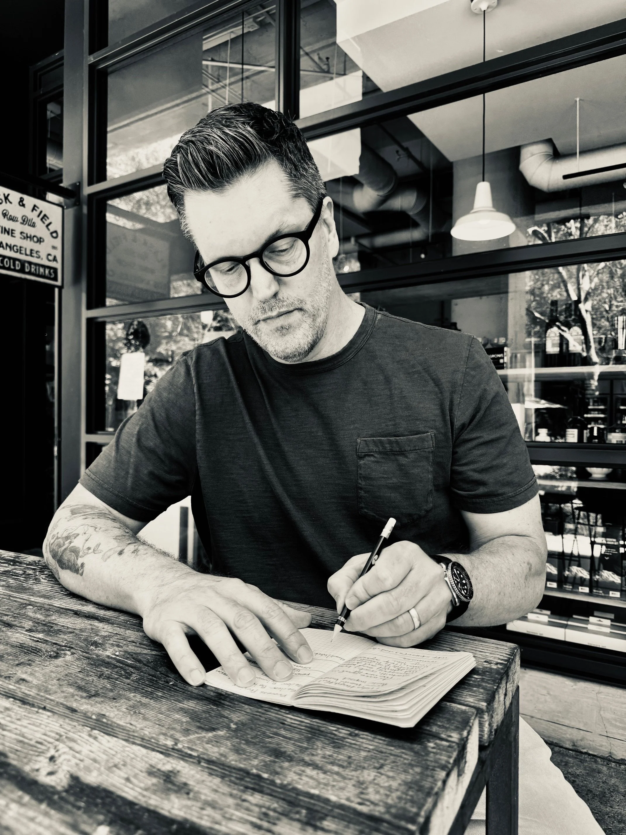 A Brad Gantt sitting at a rustic wooden table outside a modern cafe, writing in a notebook with a pen. The cafe has large windows and industrial decor.