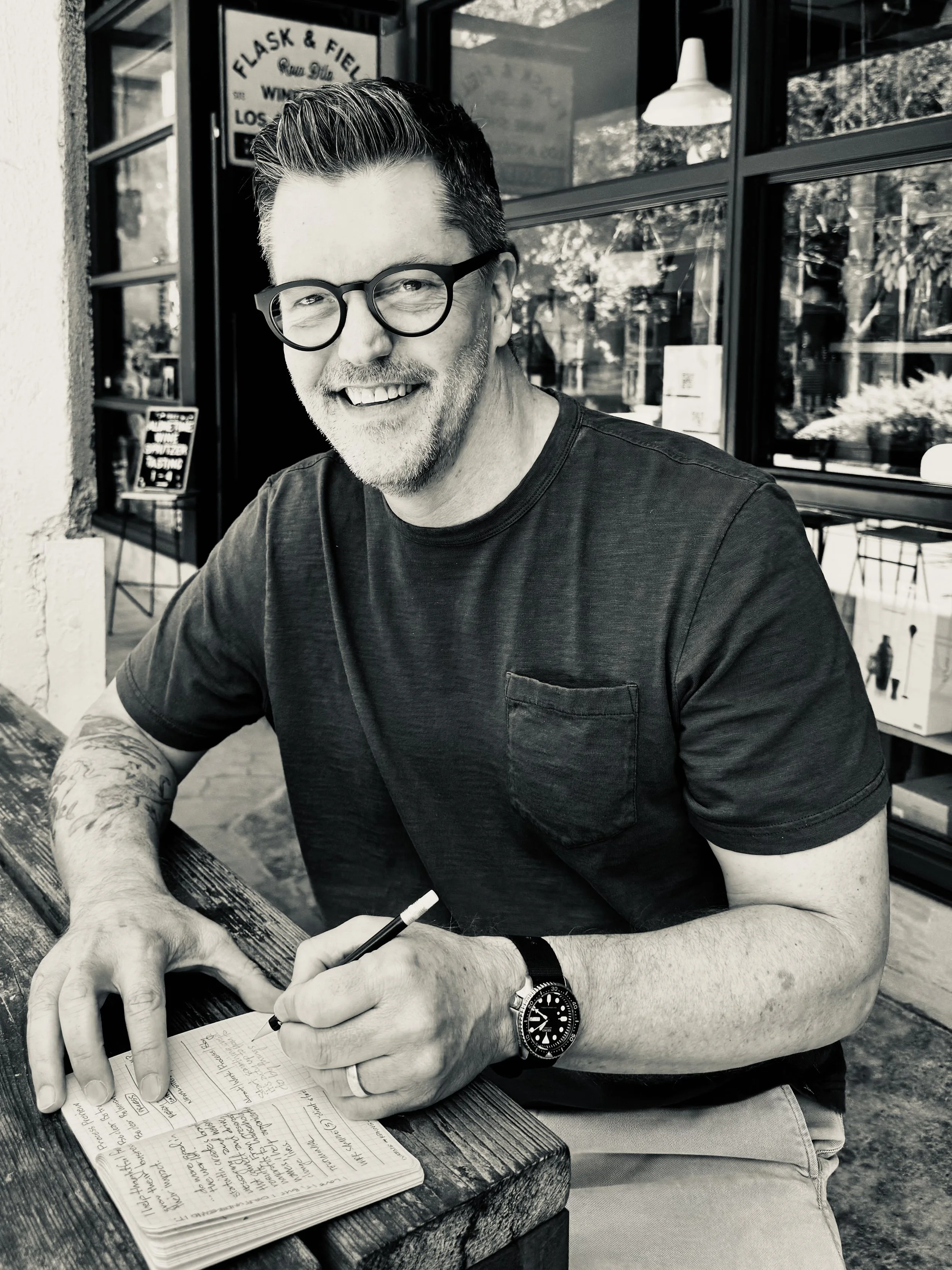 Brad Gantt, smiling, sitting at a table writing in a notebook outdoors in front of a window.