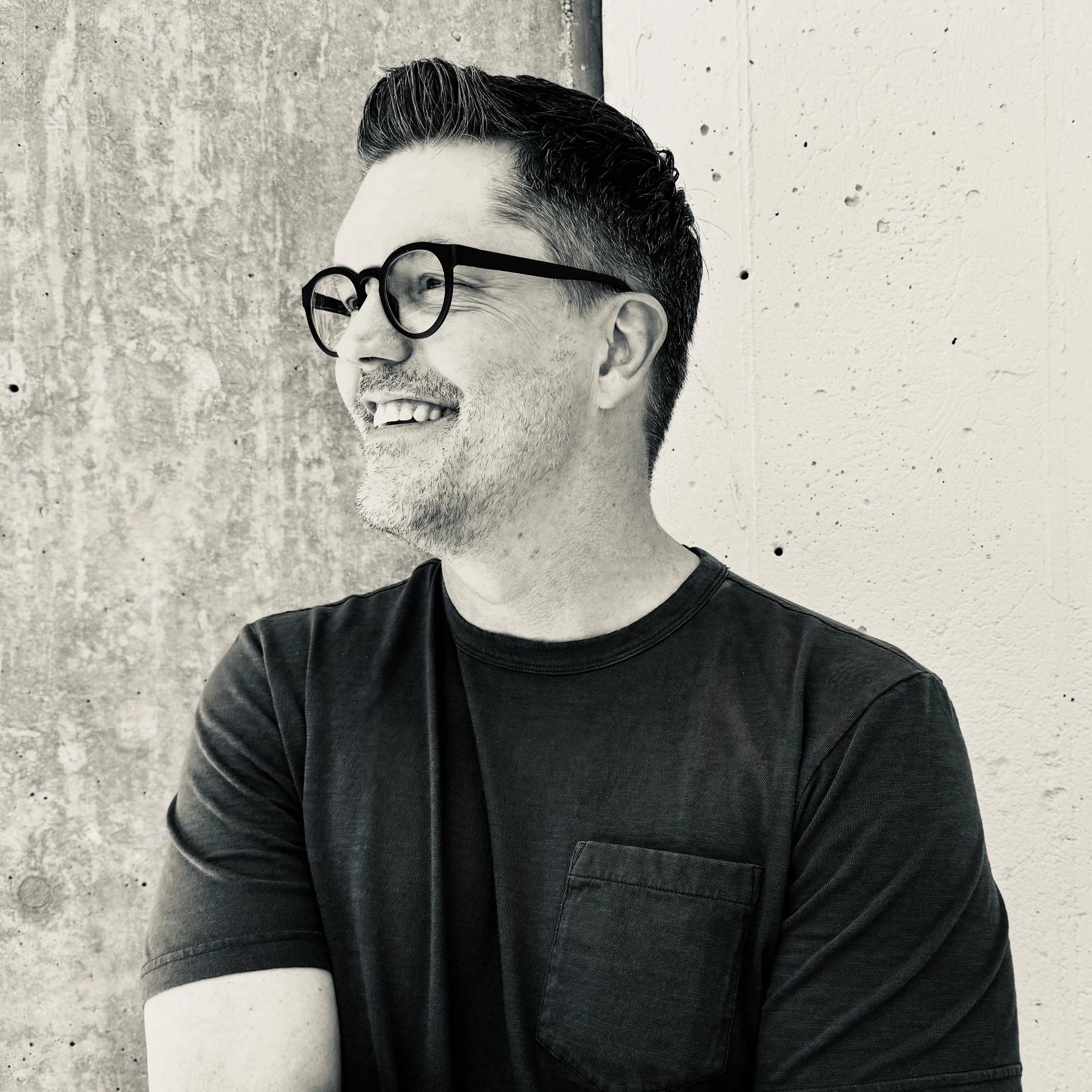 A close-up of Brad Gantt, smiling, wearing glasses and a dark T-shirt, standing against a textured concrete wall.