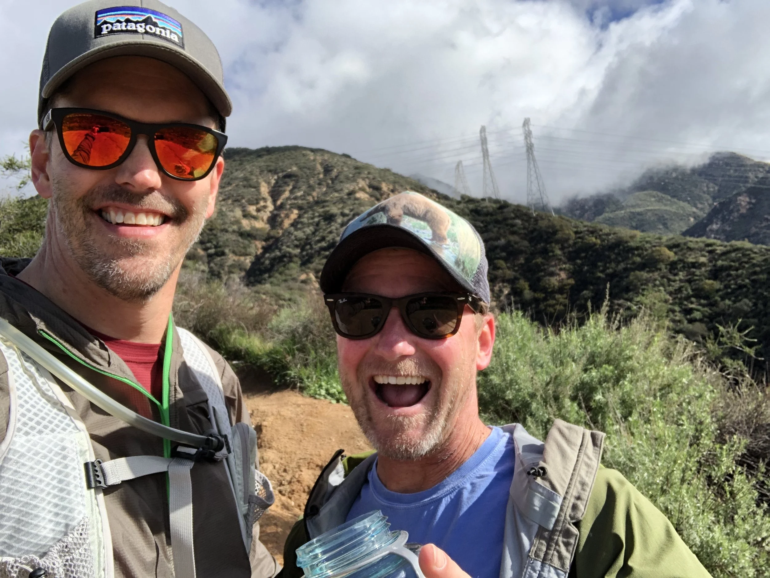 Two men smiling outdoors on a hiking trail, wearing hats and sunglasses, with mountains and power lines in the background.