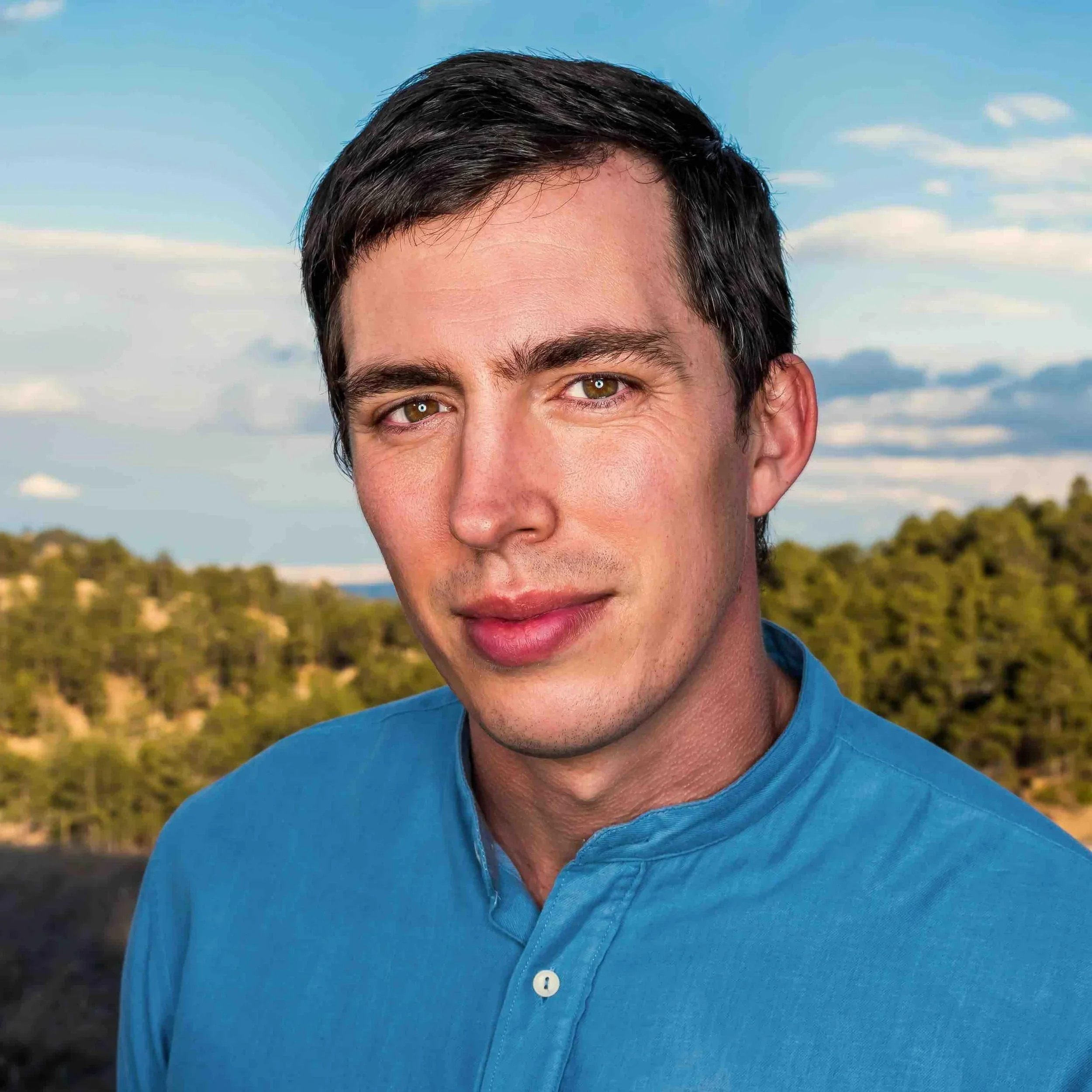 Close-up of a young man with dark brown hair, hazel eyes, wearing a blue collared shirt, outdoors with a background of trees and a cloudy sky.