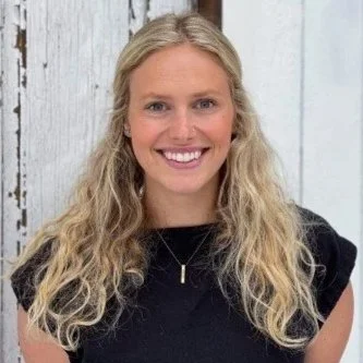 A young woman with long, curly blonde hair smiling, wearing a black top and a necklace, standing in front of a light-colored wooden background.
