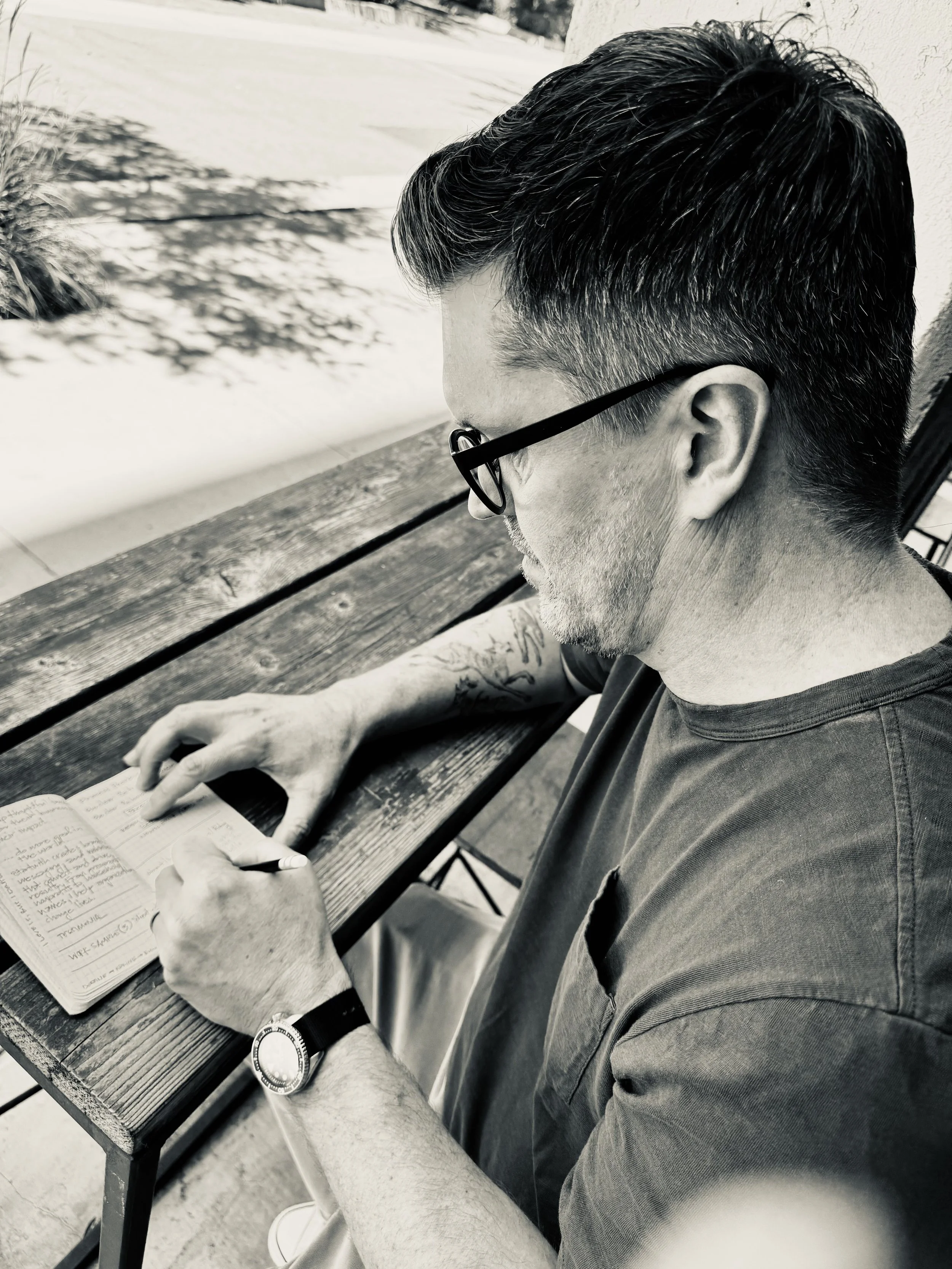 A man wearing glasses, a wristwatch, and a T-shirt is sitting at a wooden table outdoors, reading a notebook with handwritten notes and holding a pen.