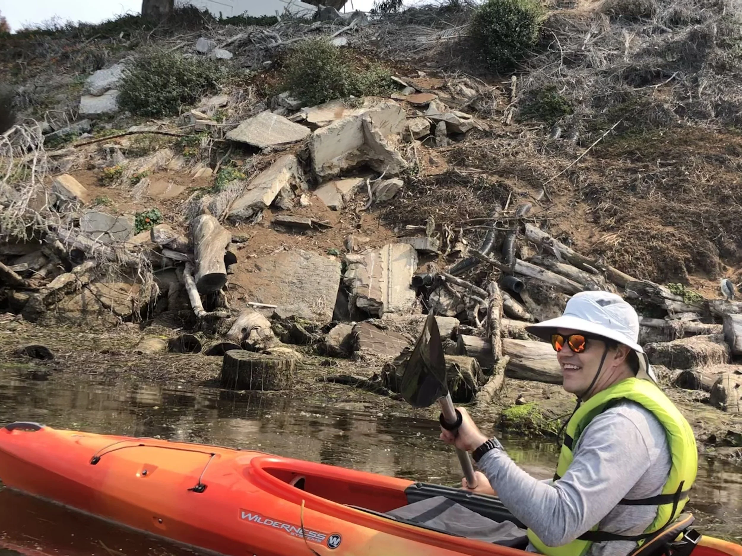 A person in a kayak on a river, smiling, wearing sunglasses and a white hat, with a background of a hillside with rocks, bushes, and dry grass.