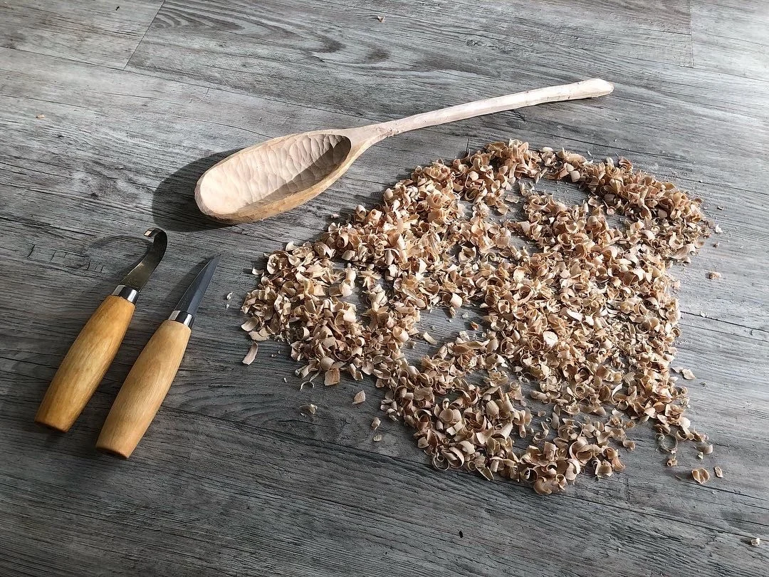 Wooden spoon, two small carving knives, and a heart shape outline made from wood shavings on a wooden surface.