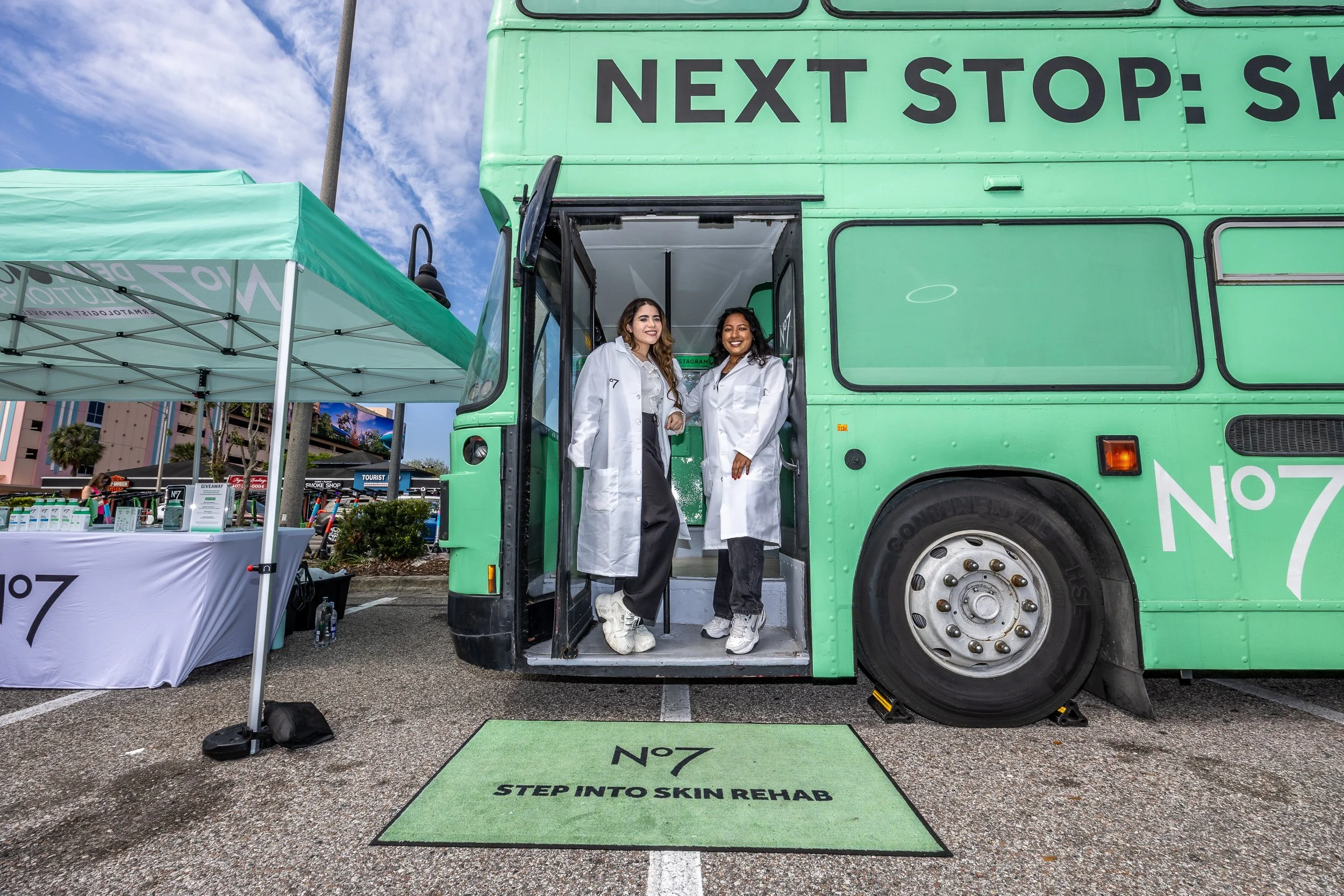 Two women standing inside the entrance of a green mobile skin rehab clinic truck, which has the words 'NEXT STOP' written on it. The women are smiling and wearing white lab coats. A green mat on the ground reads 'STEP INTO SKIN REHAB'.